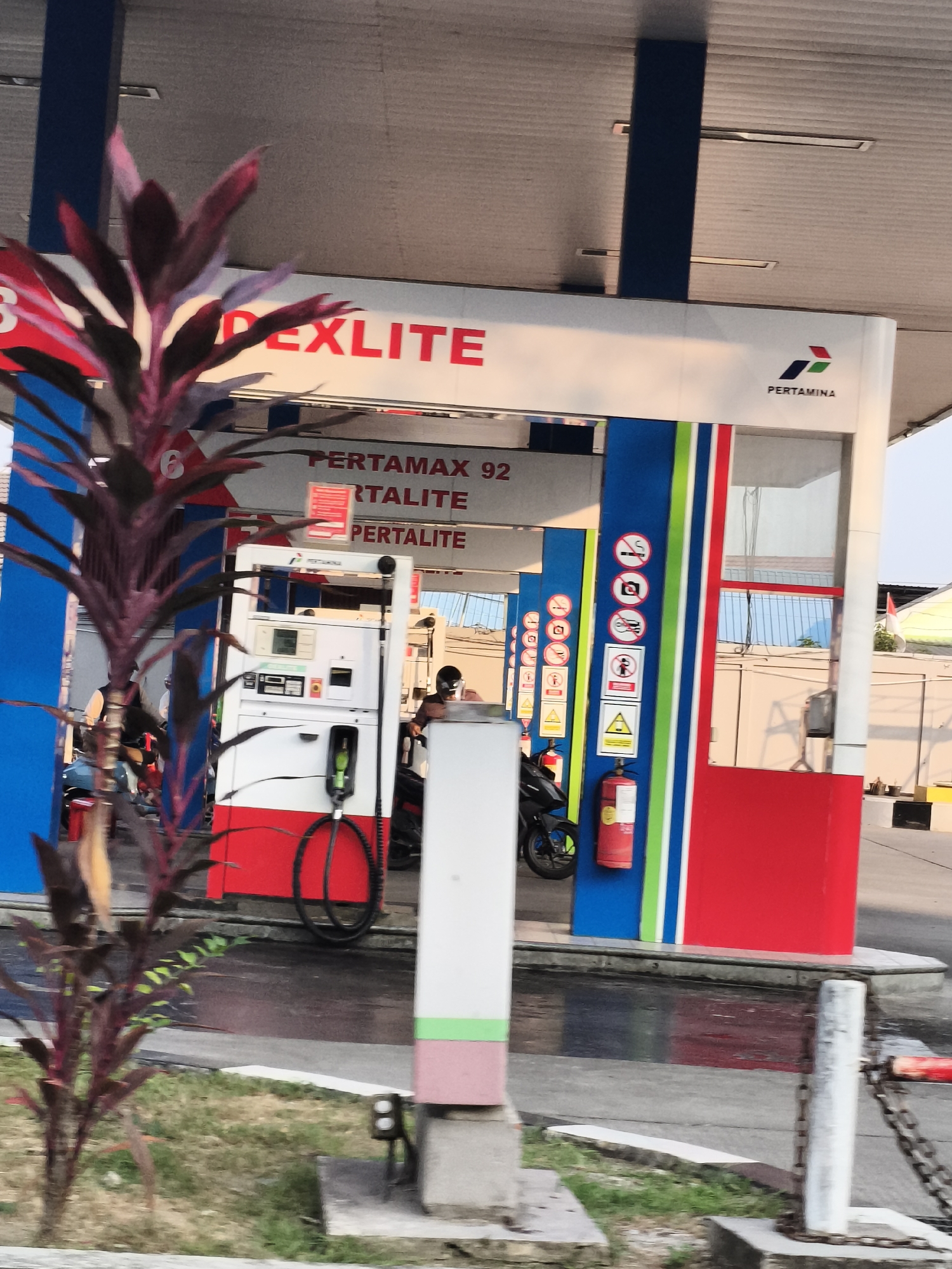A pertamina gas station in Samarinda, Indonesia is depicted. The station features multiple pumps, signage, and a canopy structure. A person on a motorbike is visible near one of the pumps. In the foreground, a potted plant with red and purple leaves is present. The ground appears wet, possibly from recent rain. The overall scene suggests a typical day at a gas station.