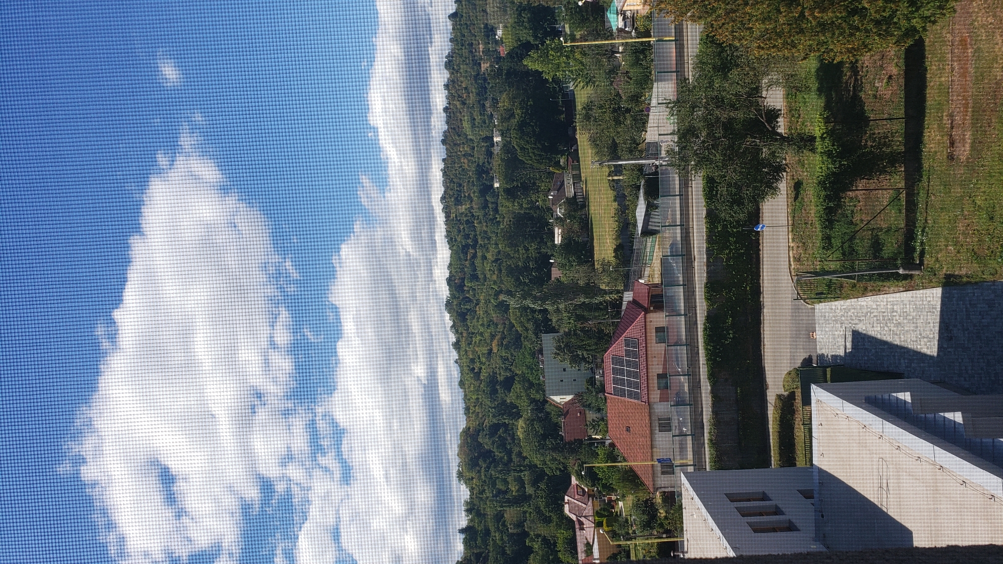 The image is an outdoor, eye-level shot of a suburban landscape in Prešov, Slovakia, as viewed through a window screen, indicated by the grid pattern overlaid on the image. The scene features a clear blue sky with scattered white clouds. In the mid-ground, a residential area is visible with several houses, one with a red roof and solar panels. Beyond the houses, a dense, tree-covered hill forms the backdrop. In the foreground, a modern building with a light-colored facade and flat roof is partially visible on the left. A paved walkway leads away from the building towards a grassy area with trees and a fence. A road runs horizontally across the mid-ground, bordered by a hedge and a fence. The overall impression is a sunny day in a mixed urban and natural environment. There are no people or animals visible in the frame.