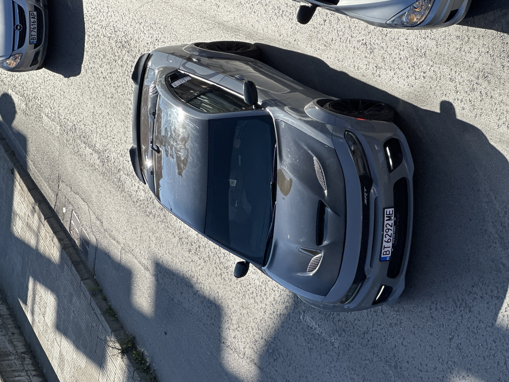 A grey Dodge Charger SRT, bearing the license plate BT 6292 ME, is parked on a sunlit asphalt road in Veliko Tarnovo, Bulgaria. The car is angled towards the viewer, with its front facing left. The car's hood has prominent vents, and a black spoiler is visible on the rear. Another silver car is partially visible to the right of the Charger. The road is lined with a concrete sidewalk and a stone wall on the left, where shadows from an unseen structure are cast. The environment appears to be a city street.