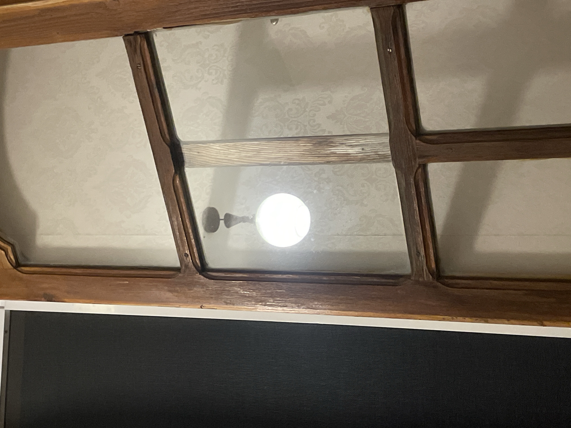 The image shows a close-up of a wooden door frame with glass panels, reflecting a light fixture with a spherical bulb. The door is set against a wall with patterned wallpaper. The left side of the image shows a dark surface, possibly a wall or another door, with a white trim separating it from the wooden door frame. The lighting suggests an indoor environment, and the reflections indicate that the photograph was taken from within a room looking outwards or through a doorway. No specific location in Tbilisi, Georgia, can be determined from the visual information alone.
