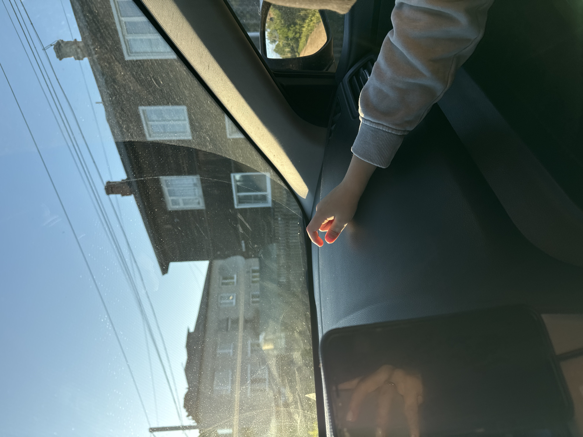 The image is taken from inside a vehicle, looking out through the windshield and side window. The scene outside depicts buildings, likely in the city of Ponaryrevo, Russia, with a bright, clear sky.
In the foreground, the dashboard of the car is visible, with a smartphone mounted on the left side. A child's hand is resting on the dashboard on the right side, illuminated by sunlight. The child is wearing a light-colored sweatshirt.
Through the windshield, two buildings are visible. The one on the right is a dark-colored structure with several windows. The building on the left appears to be lighter in color, also with multiple windows. Utility lines are strung across the sky in the background.
The side mirror reflects a dirt road winding through greenery, suggesting the car is on a road or in a rural area. The lighting indicates it is daytime, likely late afternoon due to the warm, angled sunlight. The overall impression is a snapshot of a journey through a semi-urban or rural environment.