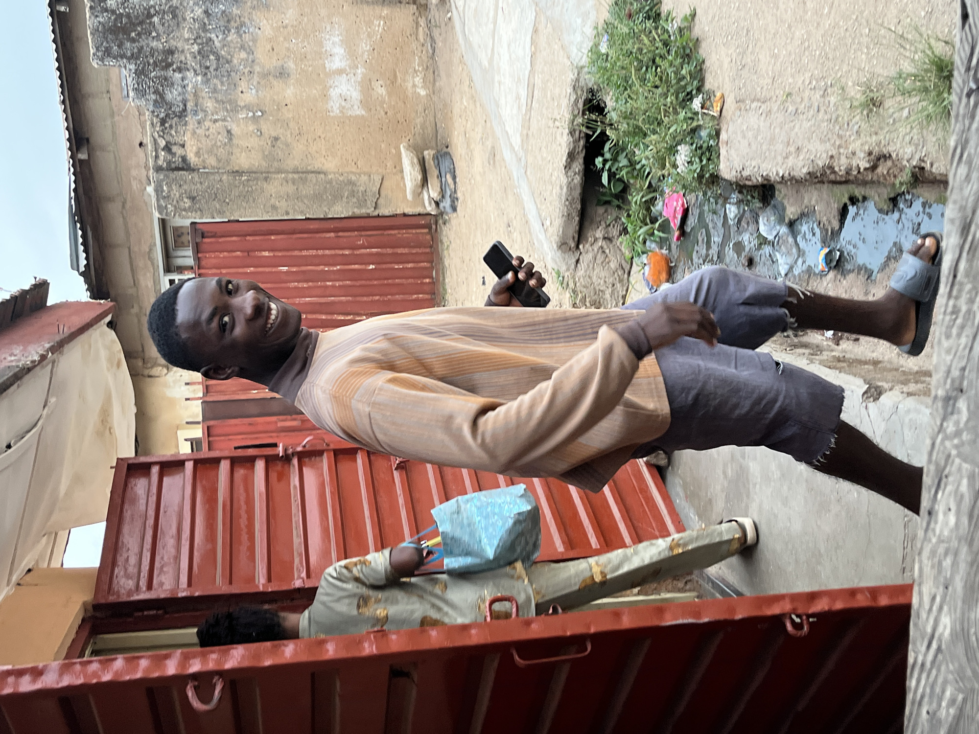 A young man stands smiling in Jos, Nigeria. He holds a phone. A person wearing a mask and carrying a blue shopping bag leans over an open stall with a corrugated red metal exterior. A drainage ditch containing plastic waste is visible behind the man. Buildings with deteriorated exteriors line the street.