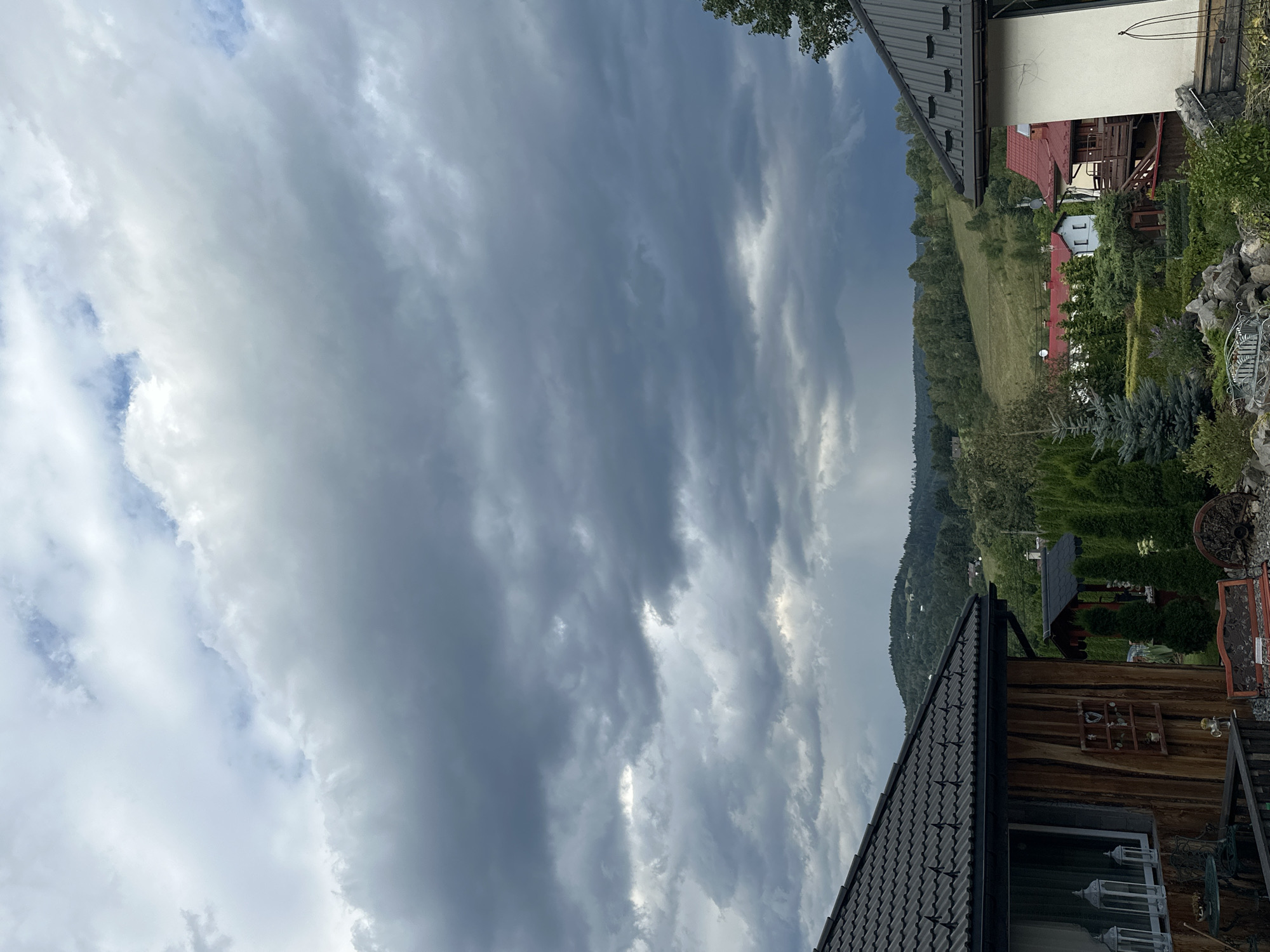 The image captures a cloudy day in Wisła, Poland. The foreground is dominated by the rooftops of two houses, one with a dark, textured roof and wooden siding, the other with a gray metal roof. A garden with manicured evergreen trees and various decorative elements is visible between the houses. In the background, rolling green hills dotted with more houses and dense forests stretch towards the horizon under a dramatic, overcast sky. Shafts of light break through the dark clouds in the distance, suggesting a possibility of rain or a clearing storm. The scene is a rural or suburban landscape, characterized by traditional European architecture nestled within a natural setting. There are no people or animals visible in the image.