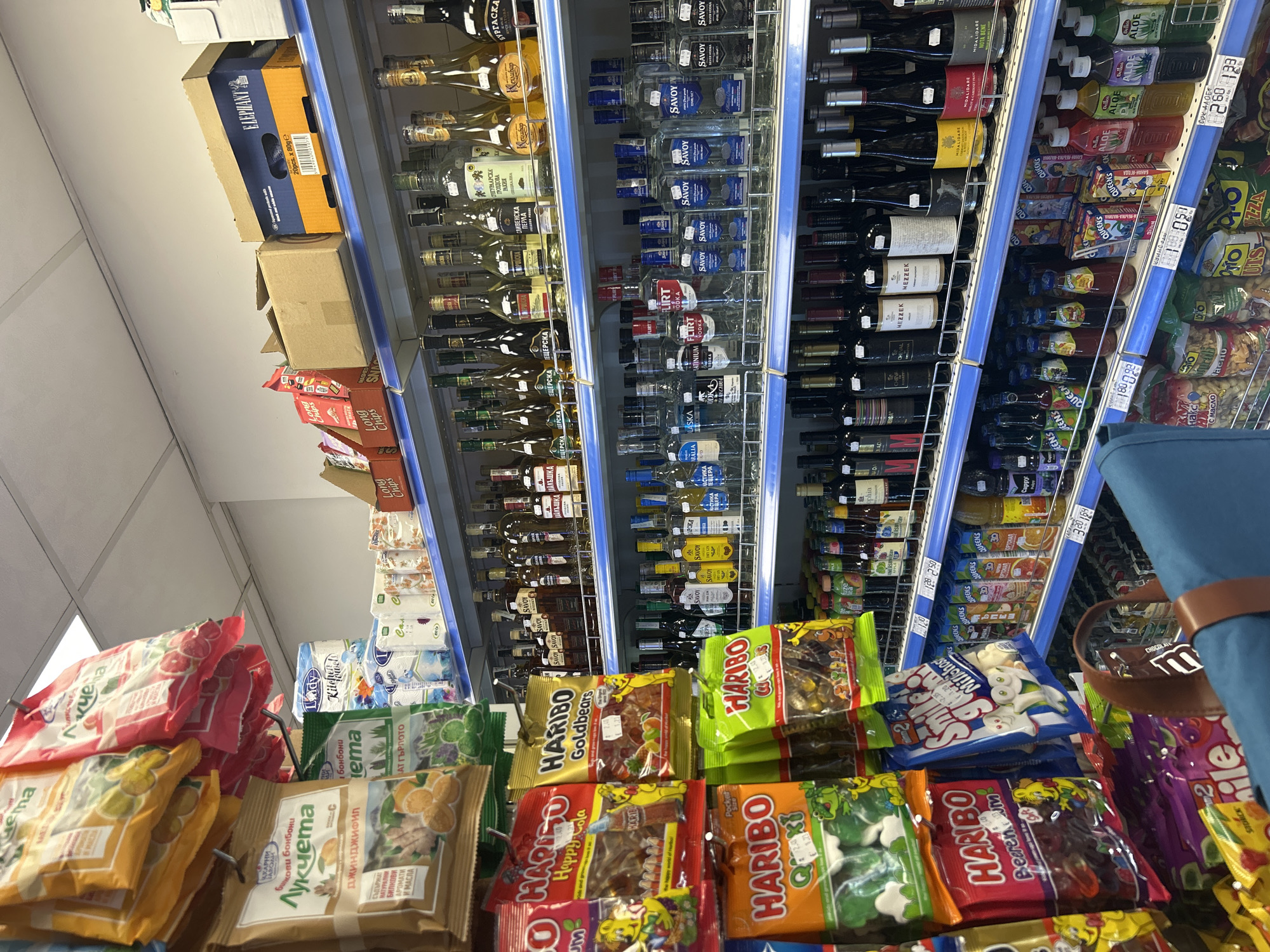 This is a vertical, eye-level shot of a grocery store aisle, likely a liquor or convenience store, with shelves stocked with various beverages and snacks. The lighting appears to be artificial, suggesting an indoor setting.
The primary subjects are the products on display. The shelves are filled with bottles of alcohol, including wine and vodka, many of which are clear glass with labels in Cyrillic script. To the right, there are also bottles of juice or other beverages, some with "ALOE" prominently displayed on their labels. Lower shelves showcase an array of packaged snacks, including gummy candies like "HARIBO" and bags of chips.
Visible text on the products includes brand names such as "SAVOY," "FLIRT," "QUEENS," "HARIBO," "ELEPHANT," and "MIDALIDARE." There are also price tags with numbers like "128 250," "260 123," and "180 092." Some text is in Cyrillic, indicating the location is likely in a Slavic-speaking region.
There are no people visible in the frame. The scene depicts a typical retail environment, with products neatly arranged on shelves, ready for purchase. The overall impression is one of abundance and variety of consumer goods. The focus is entirely on the products, with no indication of the time of day, weather, or specific emotions. The setting is utilitarian and commercial.