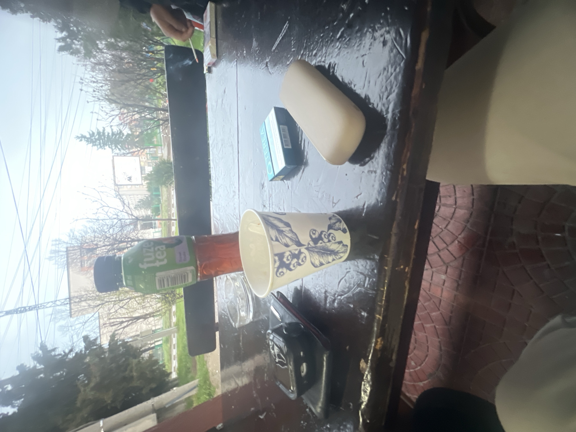 The image shows a table setting, likely outdoors, with a view of Asenovgrad, Bulgaria. A bottle of Fuze Tea, a disposable cup with a leaf pattern, a pack of cigarettes, and a white rectangular object (possibly a phone case or similar item) are on the dark-colored table. A small camera or recording device is also present. In the background, trees and a building are visible, partially obscured by the table and its contents. A person's hand holding a cigarette is visible in the upper left. The scene suggests a casual gathering.