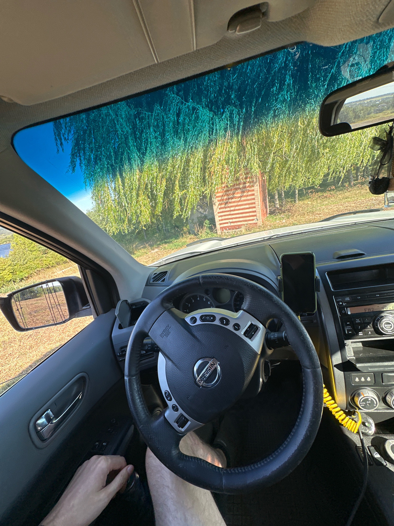 The image shows the interior of a car, likely a Nissan, viewed from the driver's perspective. The driver's hands are visible, resting on the steering wheel and near the gear shift. The car is parked outdoors, with a view through the windshield of a natural landscape featuring a large tree with hanging branches and a small red and white structure in the background. The sky is blue, and the sun is shining, casting a warm light. The dashboard of the car is visible, including the steering wheel with the Nissan logo, the car's stereo system, and a smartphone mounted on a holder. The overall impression is one of a moment of rest or observation during a car journey. The location is stated as Makiv, Ukraine, but this information cannot be visually verified from the image itself.