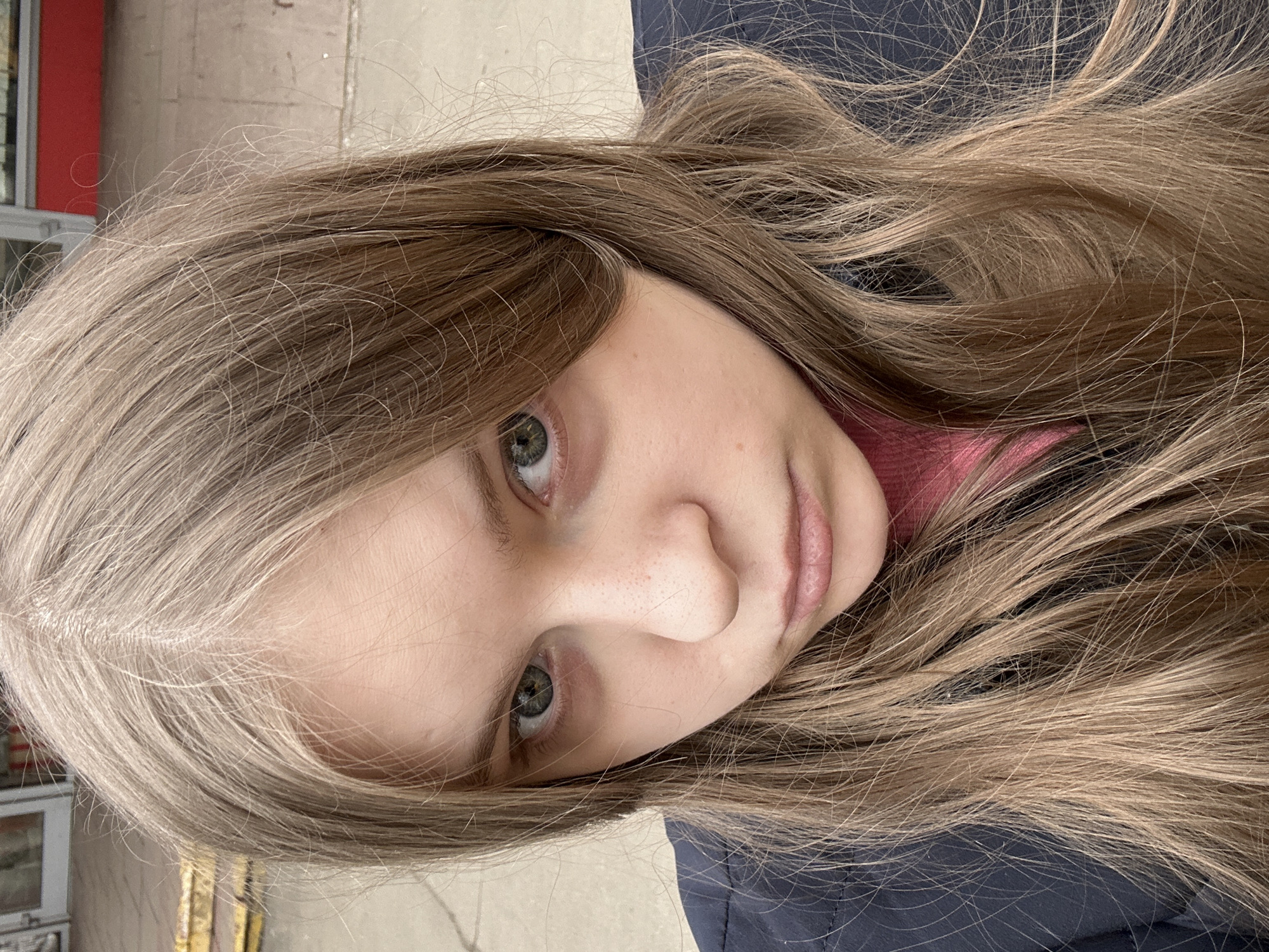 A young female with light brown hair and green eyes is pictured in a close-up shot. She is wearing a dark blue garment, and a pink top is visible underneath. The background appears to be an urban environment, potentially Moscow, Russia, based on the architectural style and the presence of a window with a red frame.