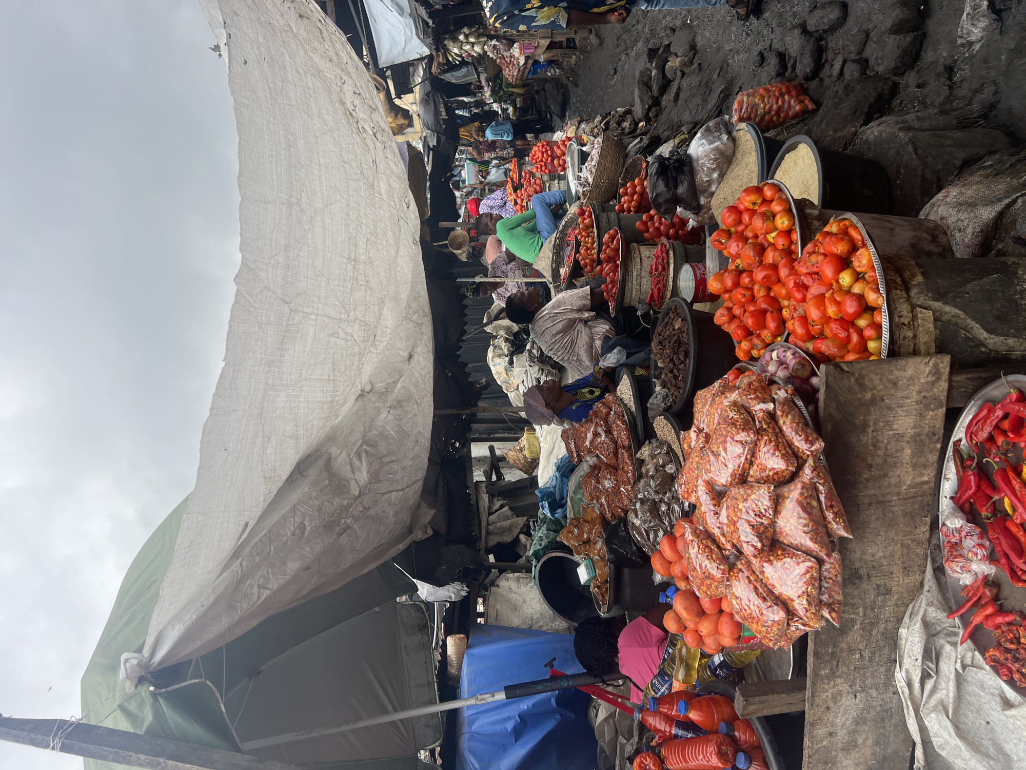 This is a street market scene in Nkpor, Nigeria, on a cloudy day. The market is characterized by makeshift stalls covered by tarpaulins, creating a sense of enclosure and shade. The ground appears to be dirt or mud, wet in places, suggesting recent rain or inadequate drainage.
In the foreground, a wooden table displays various food items. To the left, red chili peppers are piled on a metal tray and in plastic bags. Next to them are bottles of cooking oil and red sauce, as well as some whole tomatoes. A large pile of smaller red chili peppers in clear plastic bags is prominent in the center foreground. Behind this, more tomatoes are displayed in large metal bowls. There are also bags of rice and other grains in black buckets. Further back, we see more produce, including onions, and what appear to be dried goods in bulk.
Several people are present, mostly appearing to be vendors or customers. Some are seated behind their stalls, interacting with the goods. A woman in a blue and yellow patterned top is seated on the right, looking towards the market. Other individuals are visible in the background, engaged in their activities. The overall impression is one of a bustling, functional market where fresh produce and other goods are sold under somewhat rudimentary conditions. The presence of multiple tarpaulins suggests a permanent or semi-permanent market structure.