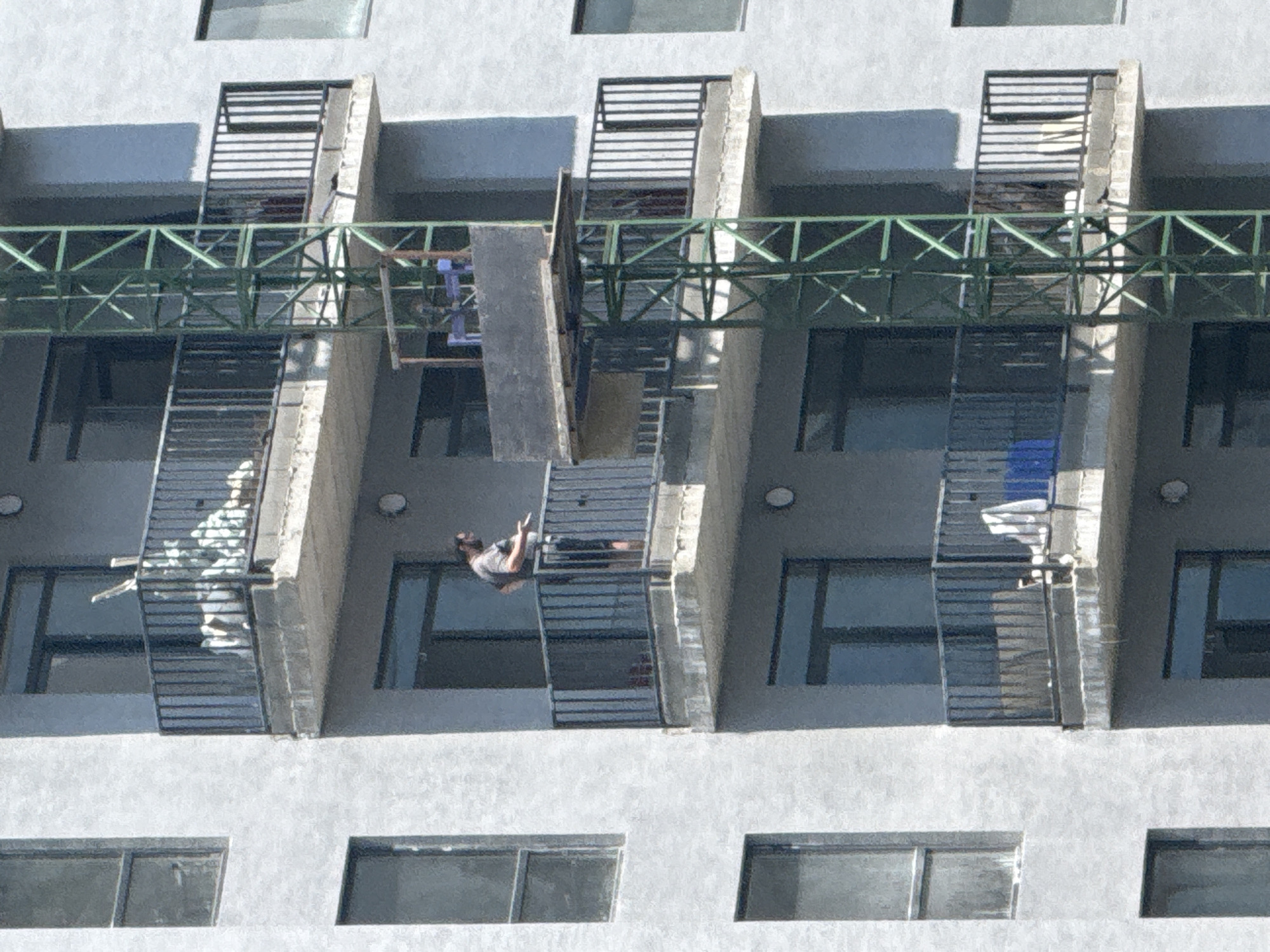 The image shows a man standing on a balcony of a multi-story building under construction. The building is a concrete structure with visible scaffolding and construction equipment, including a green metal crane. The man is wearing a grey t-shirt and black shorts and appears to be holding something in his hand. The balcony has a black metal railing and there are windows and other balconies visible on the building. The scene suggests an urban environment, possibly residential or commercial. Given the information that this image is from Tbilisi, Georgia, it depicts a typical construction site in the city.