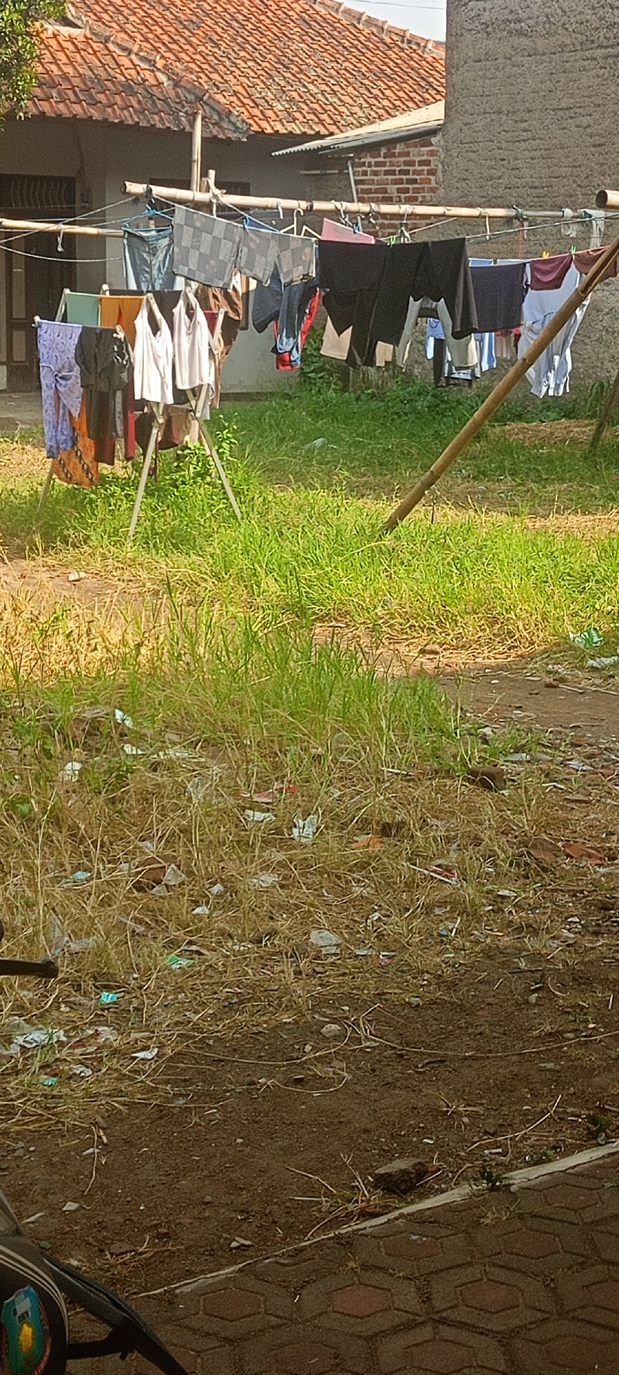 The image depicts an outdoor scene in Margahayu Selatan, Indonesia, likely a residential area. A clothesline, made of bamboo poles, is strung with various items of laundry, including shirts, pants, and what appear to be sarongs. The laundry is hung on hangers and directly on the line. The clothesline is supported by a metal drying rack and a leaning bamboo pole. In the background, a house with a red tiled roof is visible, along with a portion of a brick wall. The ground is covered in a mix of dry grass, soil, and scattered debris, including small pieces of plastic and paper. A motorcycle is partially visible in the lower left corner of the frame. The overall impression is that of a functional, everyday scene in a tropical or subtropical environment.