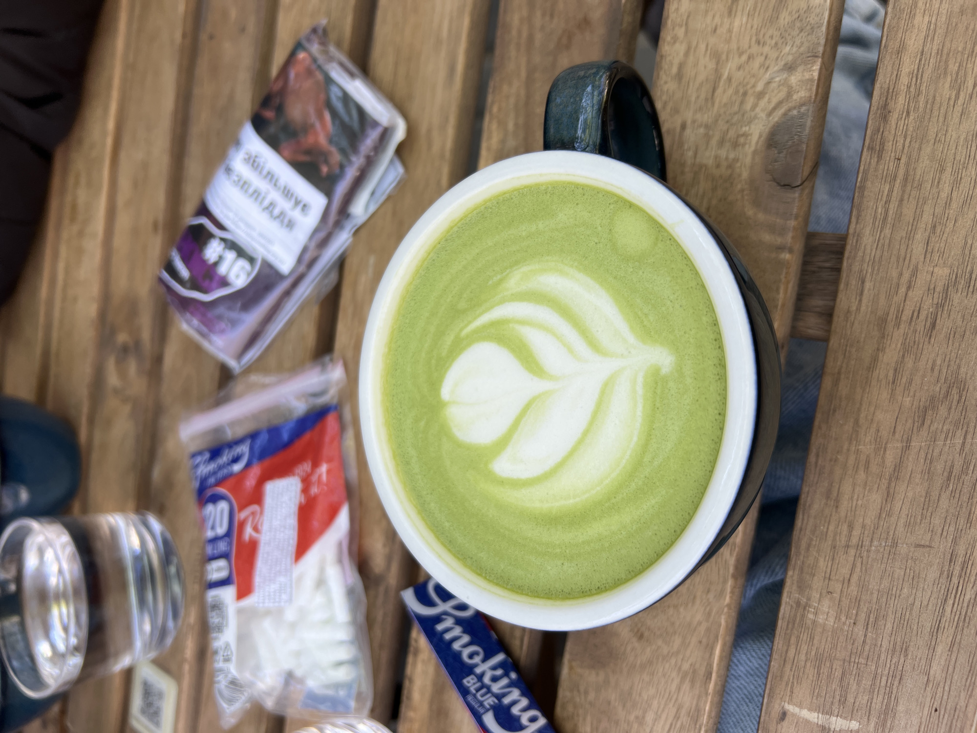 The scene captures a matcha latte on a wooden table outdoors in Odesa, Ukraine. To the left of the cup, there is a package of rolling paper, a package of cigarette filters, and a small glass pipe. The matcha latte has latte art in the center. The overall lighting suggests it is daytime. The wood texture implies it is an outdoor cafe or patio setting.