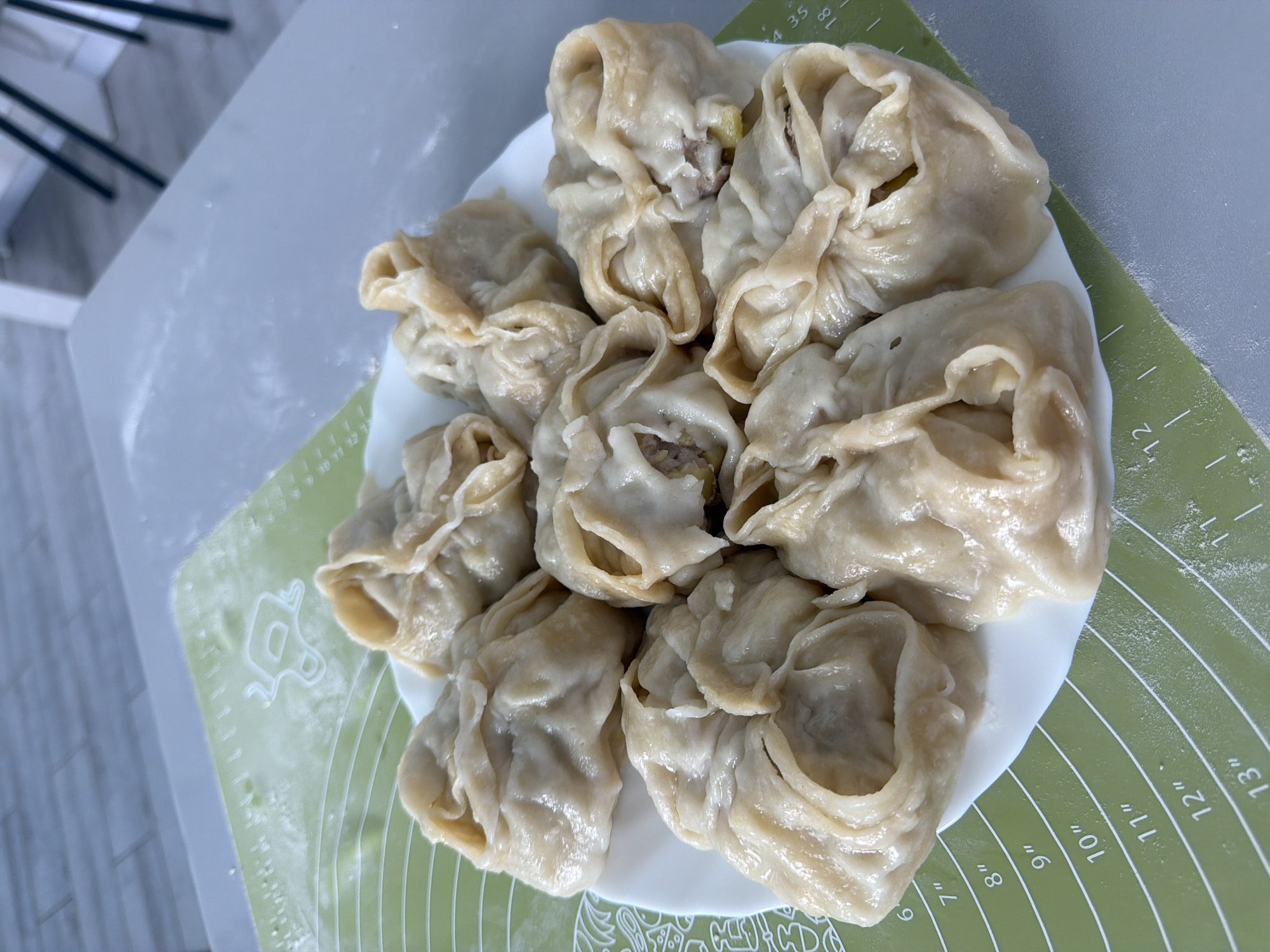 The image shows a plate of steamed dumplings, possibly manti or buuz, on a green, circular-marked pastry mat. The dumplings are beige and have a gathered appearance at the top, revealing some of the filling inside. The dumplings are arranged closely together on a white plate. The scene appears to be indoors, and the pastry mat is on a gray surface. The setting could be a kitchen or dining area. There are no people visible in the frame.
It is impossible to confirm if this image was taken in Chaikovsky, Russia.