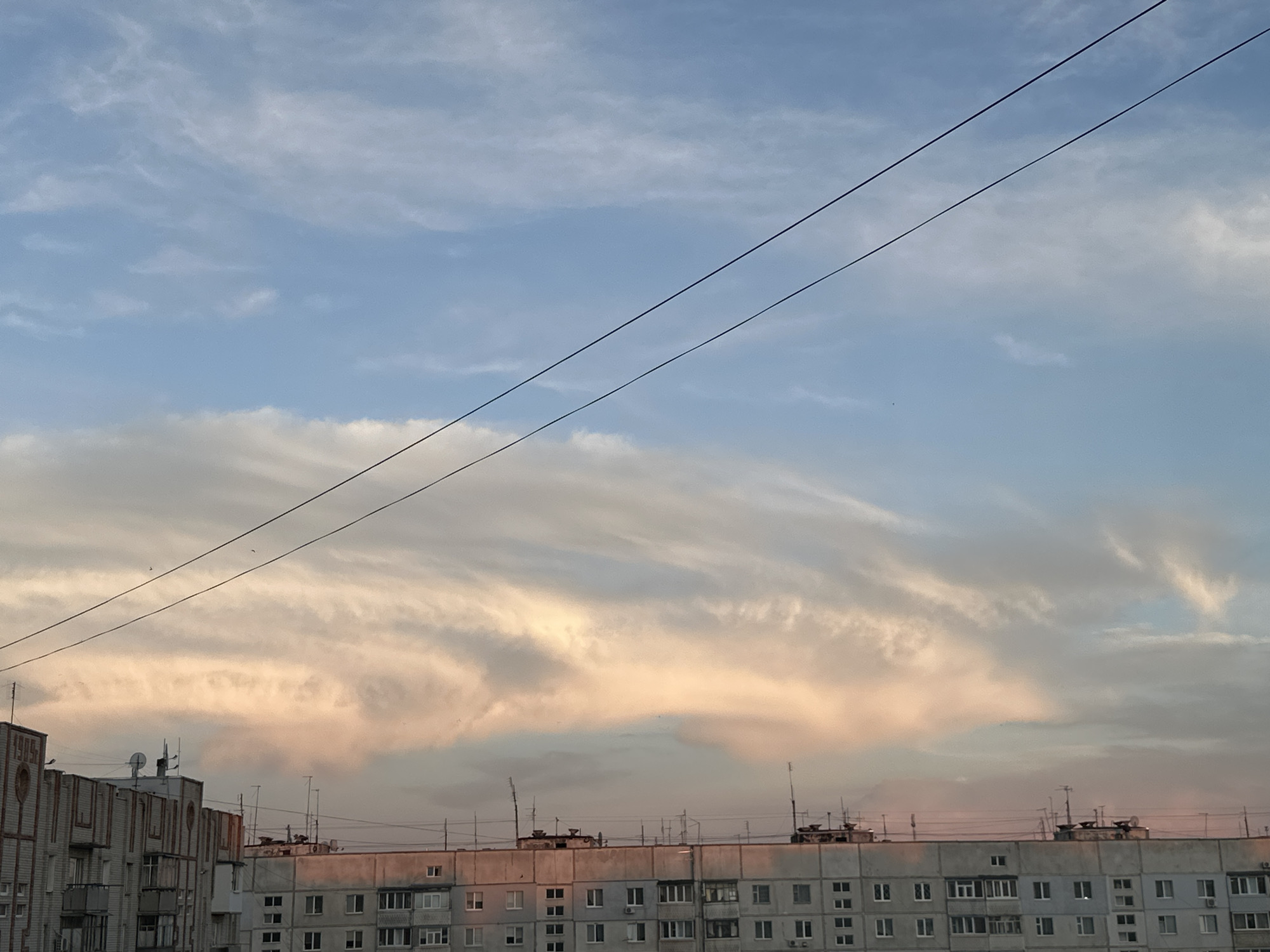 The image captures an urban landscape under a partly cloudy sky. In the foreground, the rooftops of multi-story apartment buildings are visible. These buildings appear to be constructed from concrete and feature numerous windows and satellite dishes. Overhead, two black power lines stretch diagonally across the frame. The sky is a gradient of blue and pale pink, with wispy clouds illuminated by the soft light of either dawn or dusk. The overall impression is of a residential area in a city during a transitional time of day.