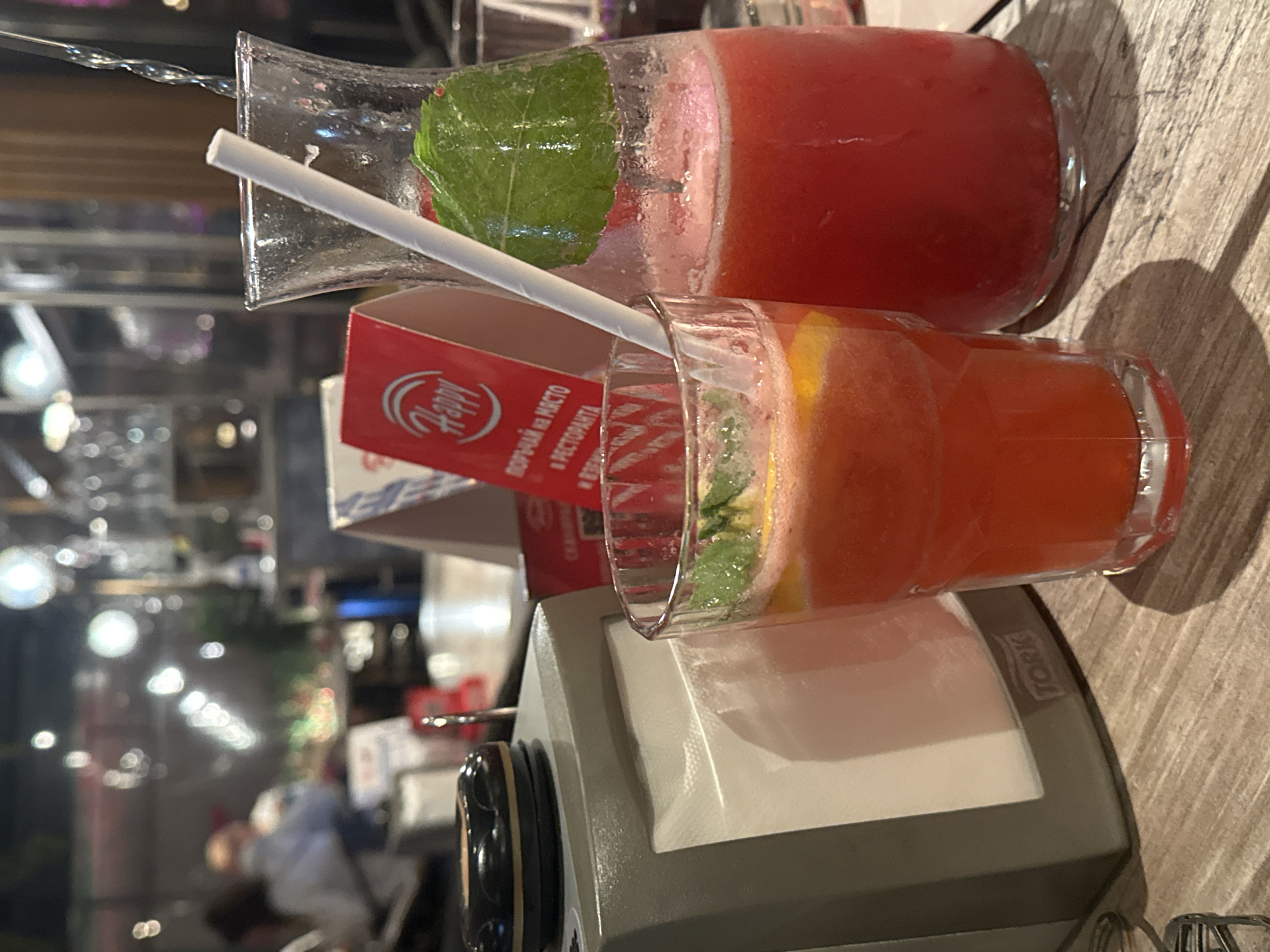 The image displays a close-up, vertically oriented shot of two refreshing beverages on a table, likely in a restaurant or bar in Sofia, Bulgaria. In the upper part of the frame, a large glass pitcher or carafe holds a red, possibly watermelon-based, drink. It is garnished with a fresh green mint leaf and has a white straw inserted. Below it, a smaller glass contains an orangish beverage, also featuring mint and slices of citrus fruit, possibly orange or grapefruit. Both glasses exhibit condensation, indicating cold temperatures.
Positioned between the two drinks is a red card with a white "Happy" logo. Visible Cyrillic text on the card reads "ПОРЪЧАЙ НА МЯСТО В РЕСТОРАНТА," which translates to "ORDER ON SITE IN THE RESTAURANT." Below the smaller glass, a grey napkin dispenser with the "TORK" brand name on its side is visible. The table surface appears to be made of wood or a wood-grain material.
The background is softly blurred with bokeh lights, suggesting an indoor evening or night-time setting, creating a relaxed ambiance. No specific individuals are clearly identifiable, but the arrangement implies a dining or social occasion where cold drinks are being enjoyed.