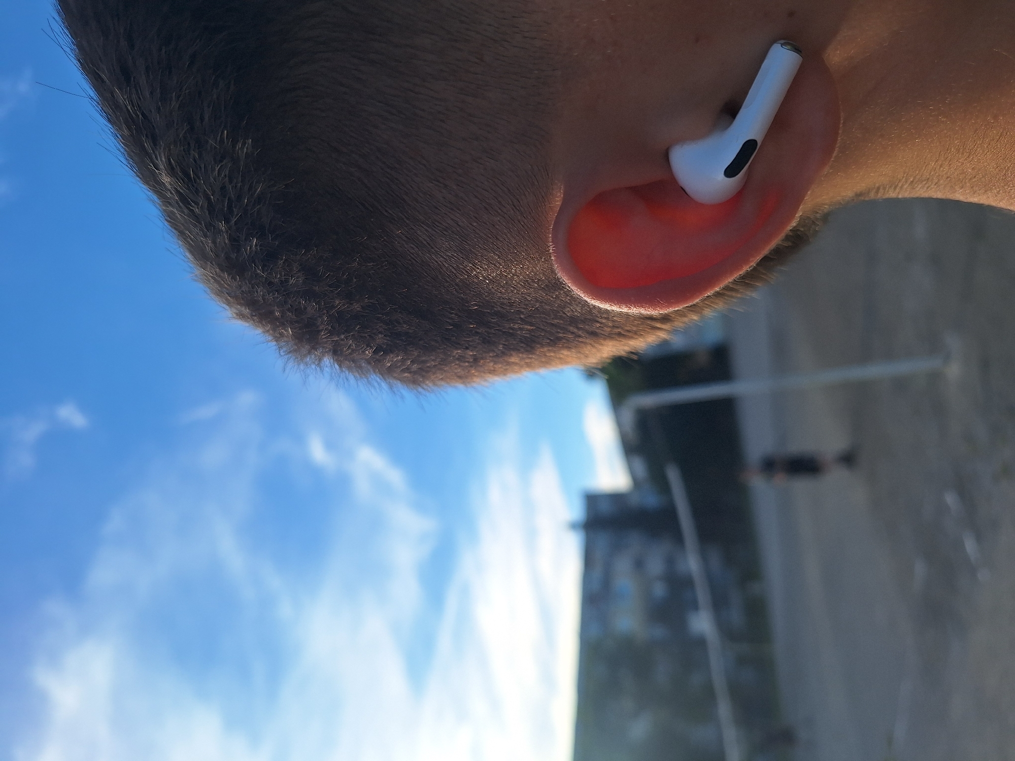 The image shows a close-up of a person's ear with a white wireless earbud inserted. The background is out of focus but appears to be an outdoor sports court, possibly in Varna, Bulgaria, with a volleyball net and a blurred figure of a person in the distance. The sky is blue with some white clouds. The lighting suggests it is daytime, possibly late afternoon due to the warm tones on the person's skin.