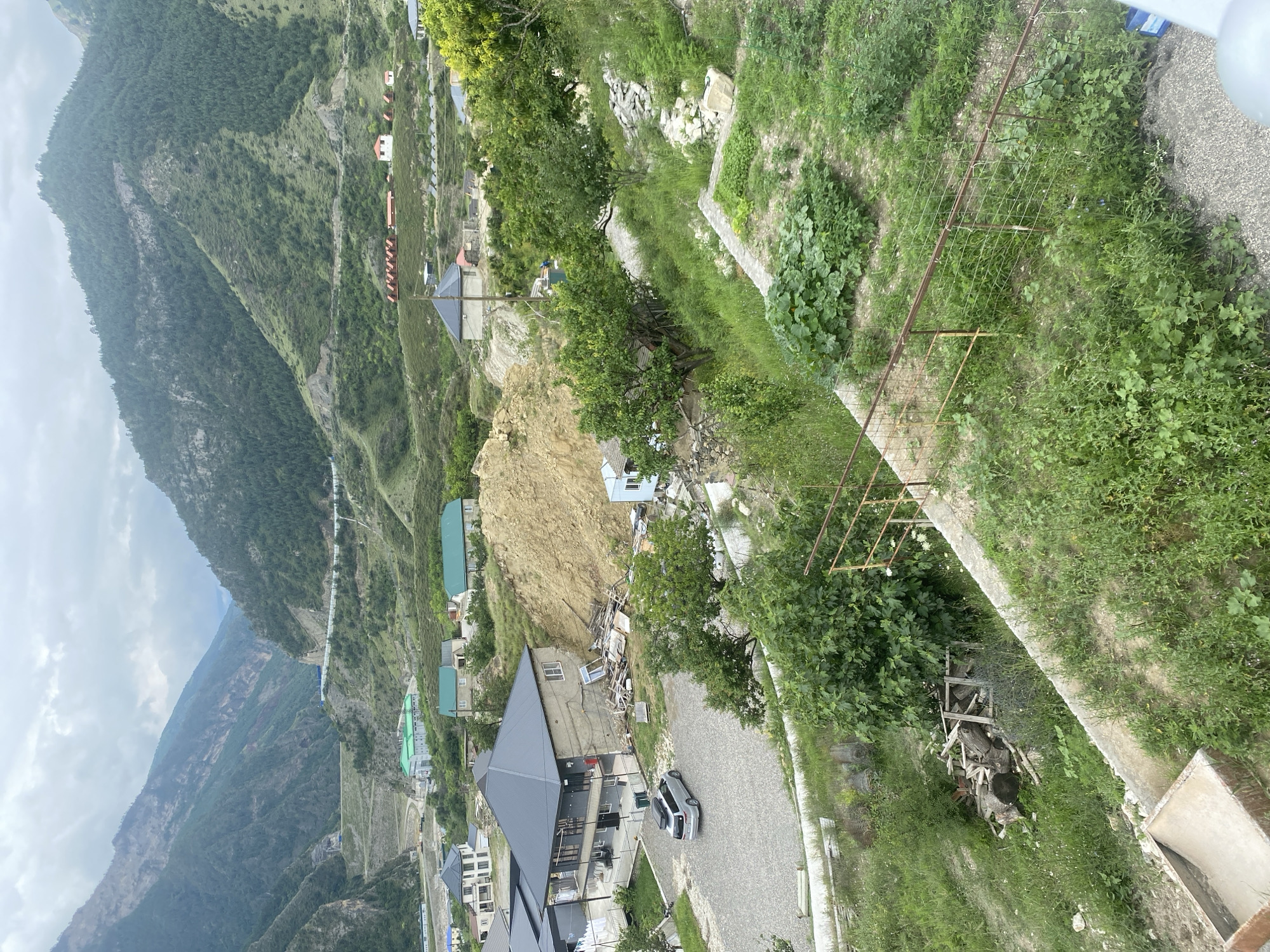 The image shows a residential area with mountains in the background. The structures visible include houses with gray and green roofs, and a few smaller buildings. A silver minivan is parked on a gravel driveway. The terrain is sloped with overgrown vegetation, including trees and garden plots. In the foreground, there is a low metal fence and some piled lumber. The sky is overcast. The scene depicts a rural or semi-rural environment.