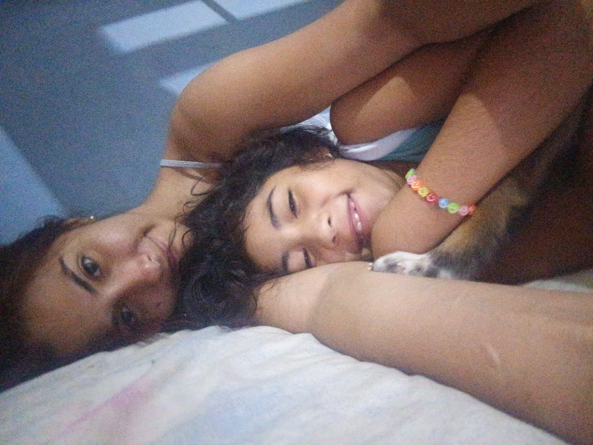 A young girl lies on a white bed next to a woman. The girl has dark curly hair and is wearing a colorful beaded bracelet. A dog is partially visible, with a paw resting on the girl's arm. The woman has dark hair and eyes. The background includes a blue wall and a shadow. The location is not explicitly discernible in the image, and cannot be inferred to be Juan Griego, Venezuela.