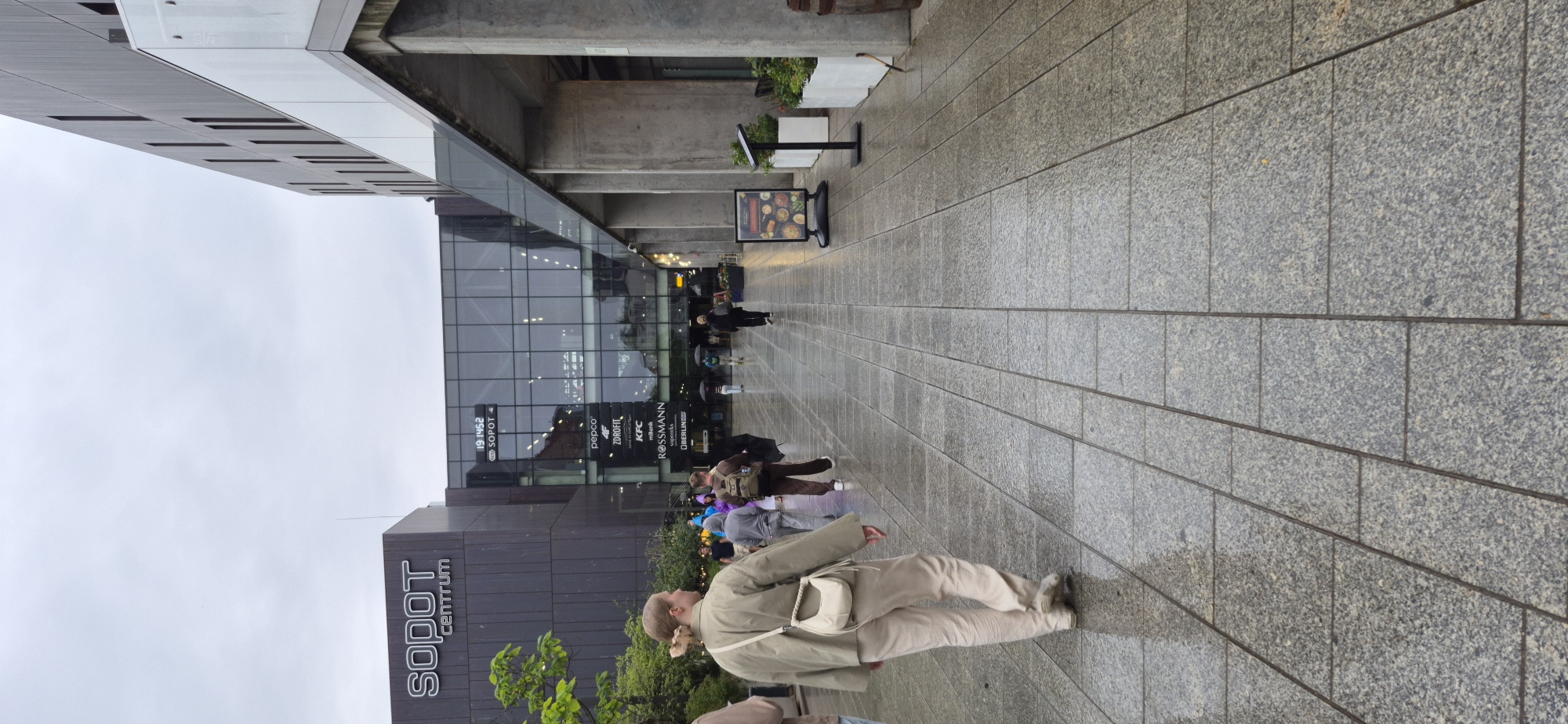 The image depicts an exterior view of the Sopot Centrum shopping mall in Sopot, Poland. The scene is captured on a rainy day, evident from the wet pavement tiles that reflect the overcast sky and surrounding architecture.
In the foreground, a person walks away from the camera, wearing a light olive-green jacket, cream-colored pants, and a small cream shoulder bag. The wet, grey granite tiles of the walkway are clearly visible and patterned.
Further into the scene, several other individuals are walking in various directions. Some are carrying umbrellas, reinforcing the rainy conditions. The Sopot Centrum building features a modern architectural design with a large glass facade that showcases several retail brand logos, including "pepco," "ZDROFIT," "KFC," "mBank," "ROSSMANN," and "OBERLIN." Above the entrance, a digital display shows the time "19:45:52" and "SOPOT".
To the right of the walkway, there are concrete columns supporting an elevated structure, and a sign advertising food is displayed on a stand. Planters with greenery are also visible. The sky is uniformly grey, indicating a cloudy or stormy weather. The overall atmosphere of the image is one of a typical urban environment on a wet day, with people going about their activities despite the weather.