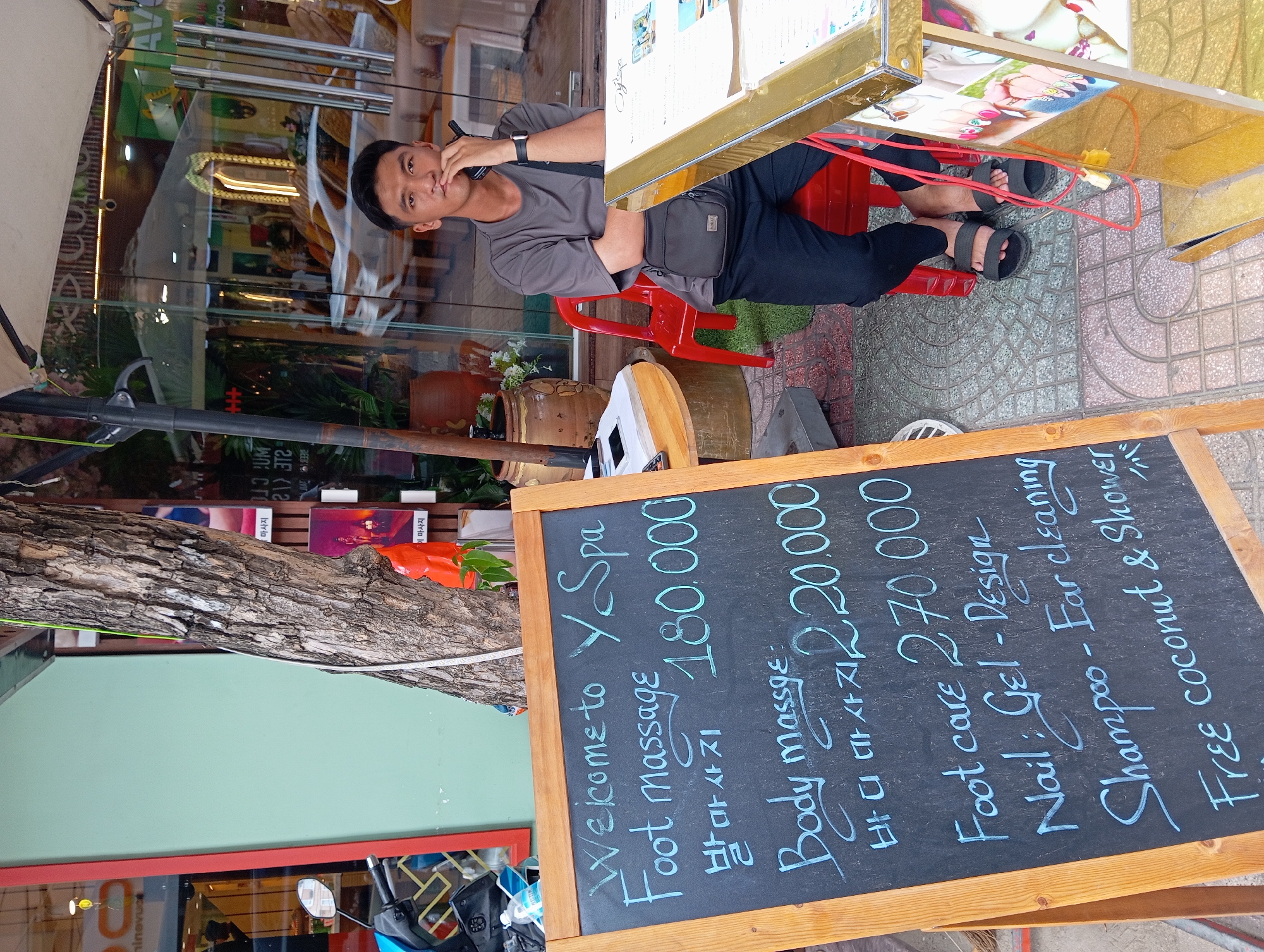 In Nha Trang, Vietnam, a sign on a chalkboard stands near the entrance of "Y Spa," offering a menu of services. The sign, written in blue chalk, lists "Welcome to Y Spa," "Foot massage 180.000," "Body massage 220.000," "Foot care 270.000," "Nail: Gel - Design," "Shampoo - Ear cleaning," and "Free coconut & Shower." Below the English text for foot and body massage, Korean characters are present. A young man, seated on a red plastic chair, is positioned behind a counter, looking directly at the camera. He is wearing a grey t-shirt and black pants, with a black crossbody bag. He holds a black object to his mouth, possibly a phone or a cigarette. The background reveals the interior of the spa with glass doors and windows, reflecting a white car and interior furnishings. A tree trunk with rough bark is to the left of the sign, and a blue motorcycle is partially visible in the background. The ground is paved with decorative tiles.