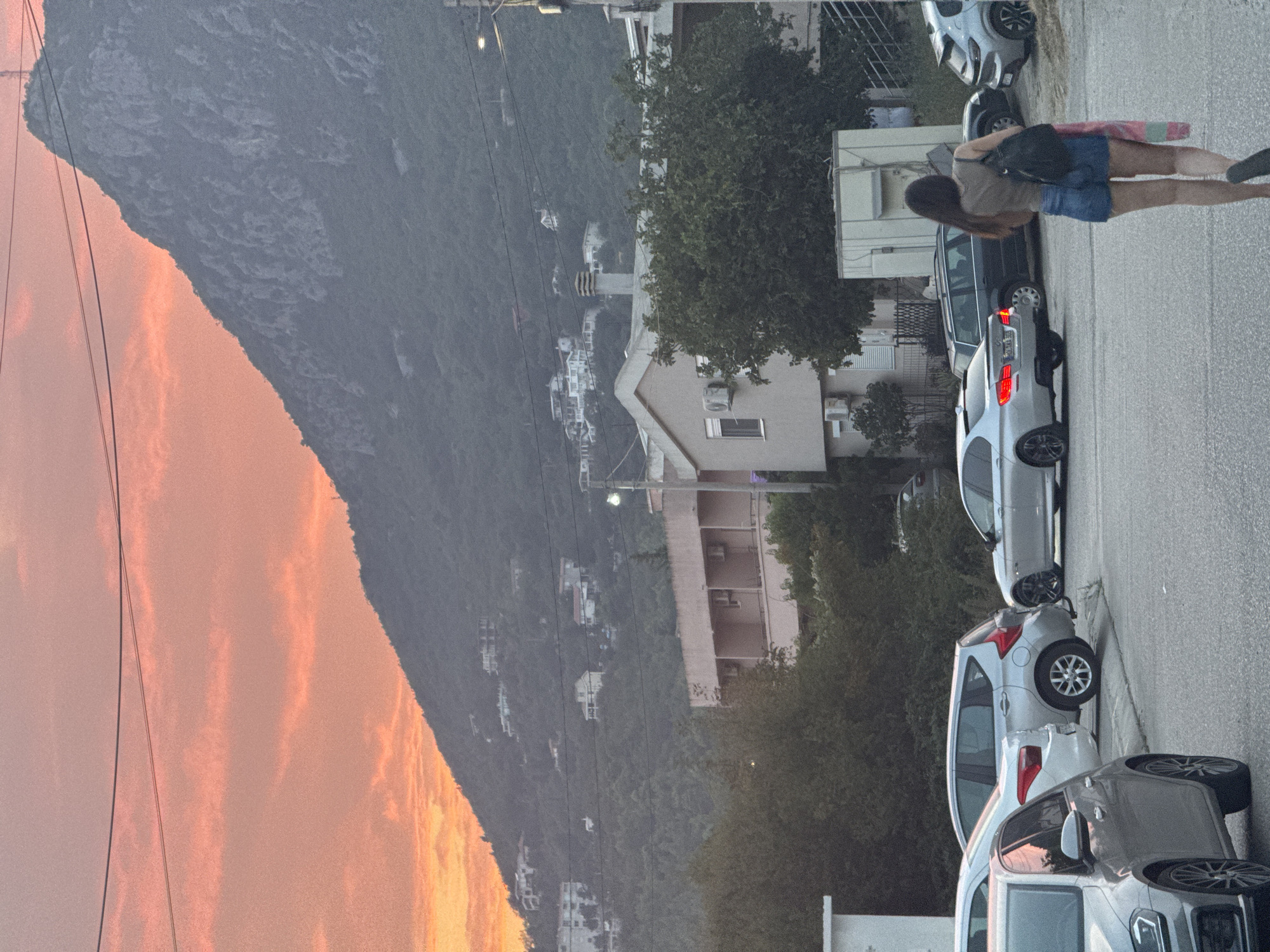 In Sutomore, Montenegro, a woman walks away from the camera on a paved road during sunset. She is wearing a taupe tank top, denim shorts, and carrying a black backpack over her left shoulder, with a striped cloth draped over it. A silver sedan is parked to her right. In the background, several other cars are parked along the street, including silver, grey, and black vehicles. A multi-story building with a light pink facade and balconies is visible behind the parked cars, partially obscured by trees. Further in the background, a steep, tree-covered mountain rises sharply, its peak silhouetted against a vibrant orange and pink sky. Houses are scattered across the mountainside. Overhead, power lines traverse the frame.