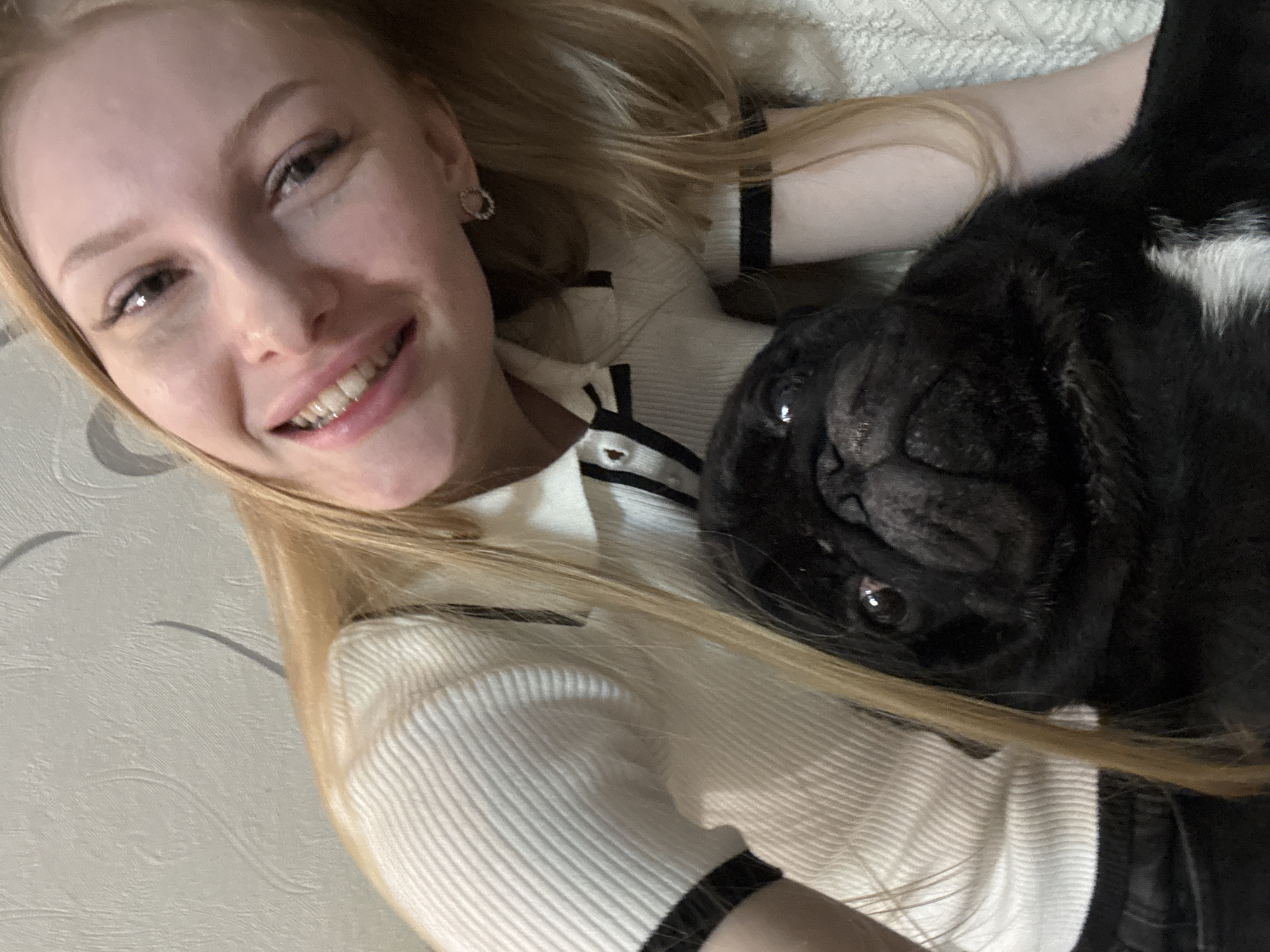 A light-skinned young woman with long blonde hair is lying down, smiling at the camera. She is wearing a white ribbed sweater with black trim. To her right is a black pug dog with white markings on its chest, lying on its back, its face close to the camera. The background appears to be a textured surface, possibly a bed or couch. This photo was taken in Perm, Russia.