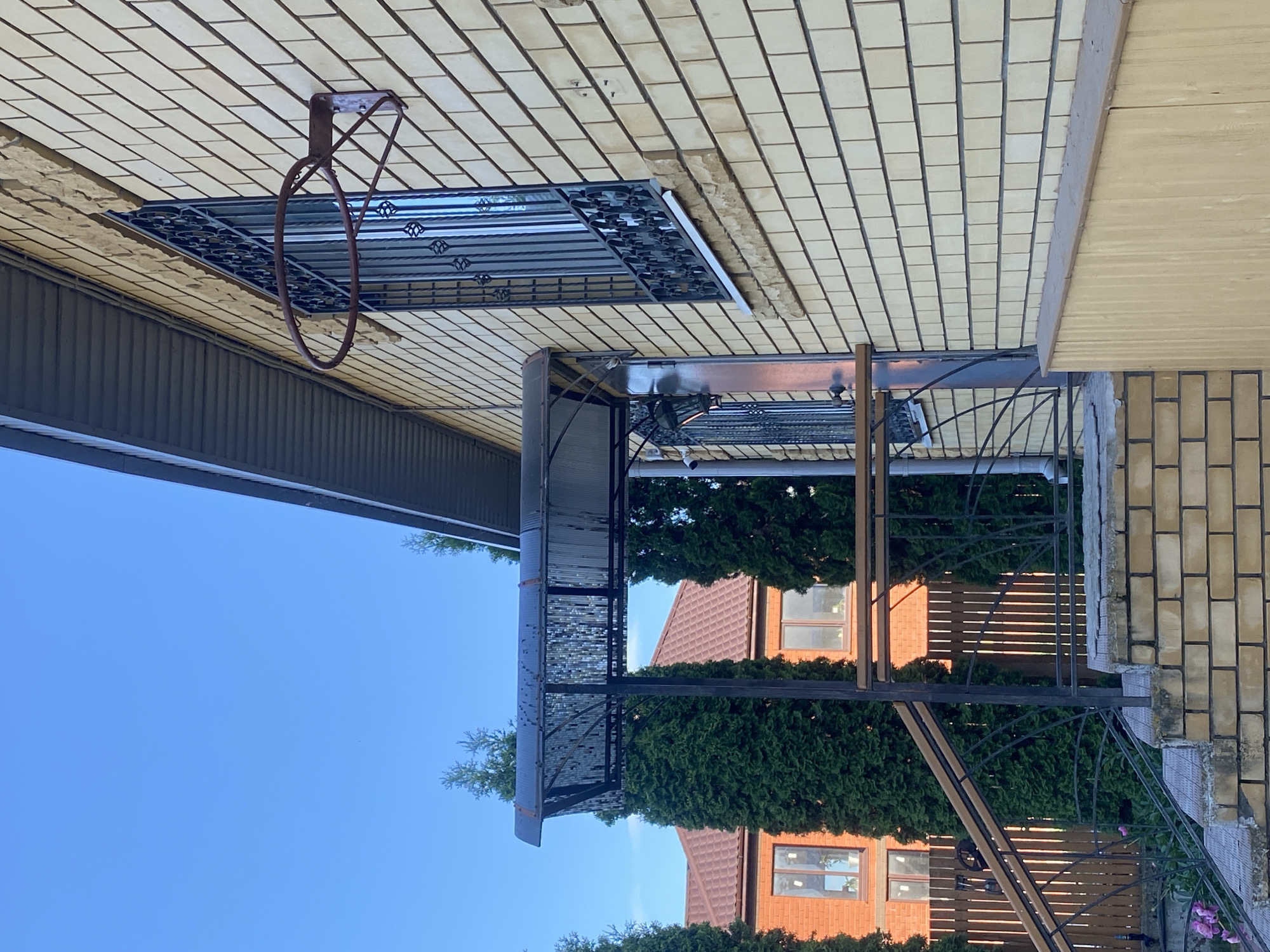 A photograph taken at a low angle shows the side of a light yellow brick house with a dark grey, ribbed roof overhang. A rusty basketball hoop is attached to the brick wall above a window with black wrought-iron bars. To the left, a covered porch with a patterned translucent roof is supported by dark posts. A metal railing with wooden accents leads up a brick staircase. Several tall, green cypress trees are planted in front of the house and a brown-tiled roof house is visible behind them. The sky is clear and blue. The location is Volzhsk, Russia.