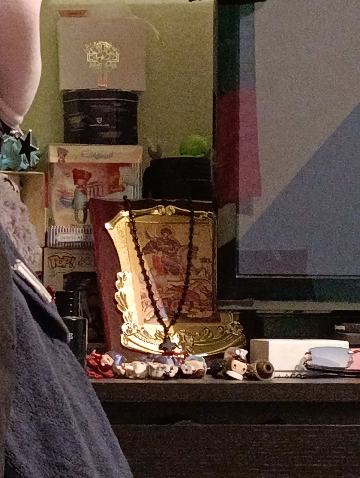 A cluttered shelf holds various objects including an ornate gold-framed religious icon with a rosary draped over it, assorted small figures, a white rectangular object, and a pair of glasses. Behind these, there are stacked boxes with printed designs and a black tin. A monitor is visible on the right. The location, Emmerich on the Rhine, Germany, cannot be confirmed from the image alone. There are no people visible. The environment appears to be an interior setting.