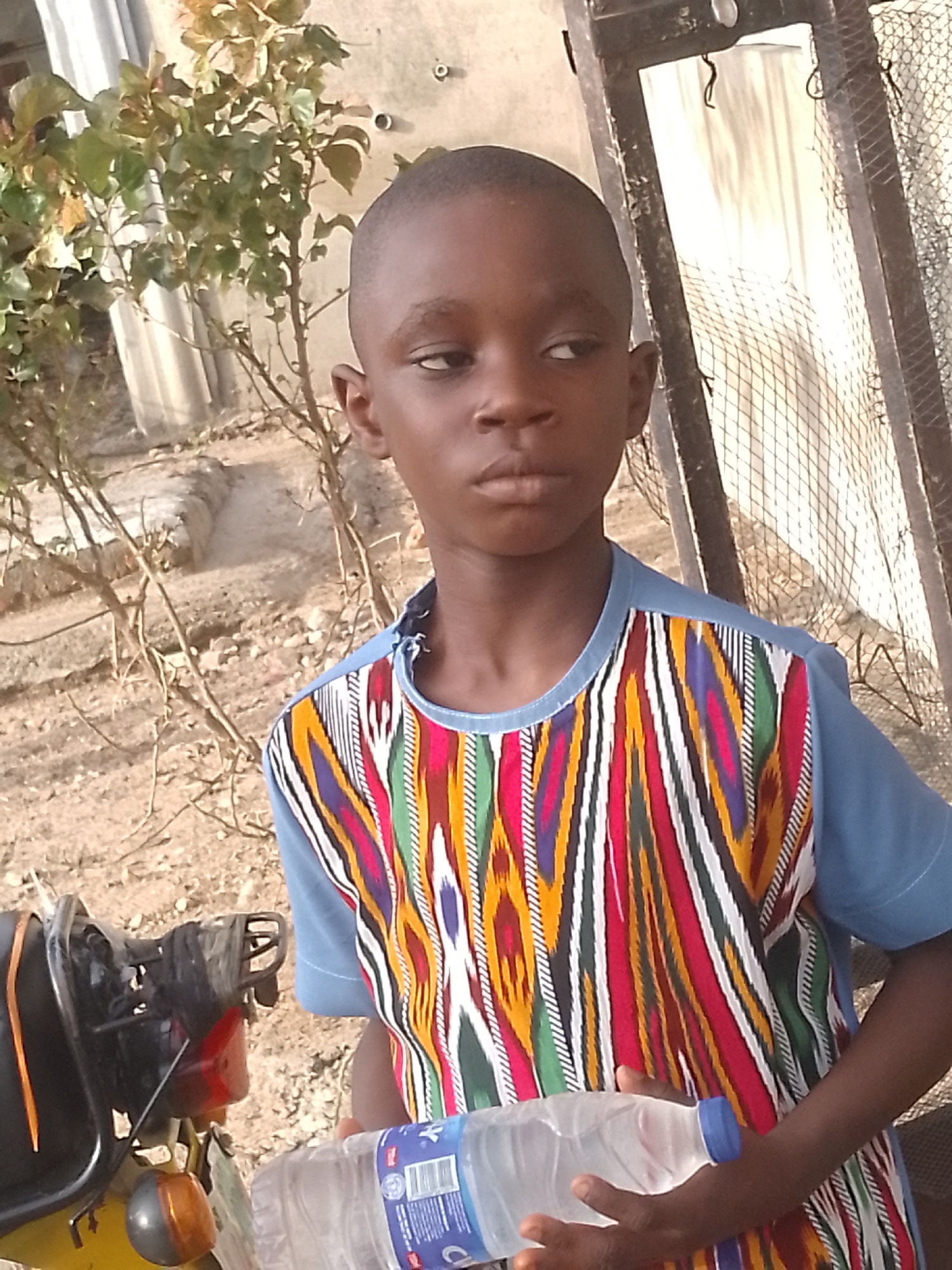 The image depicts a young boy standing outdoors in Unknown, Unknown. He has a closely shaved head and is wearing a colorful, patterned shirt with short blue sleeves. He is holding a water bottle. To his left is a motorcycle. Behind him, there is vegetation and a fence. The area appears to be a yard or outdoor space near buildings.