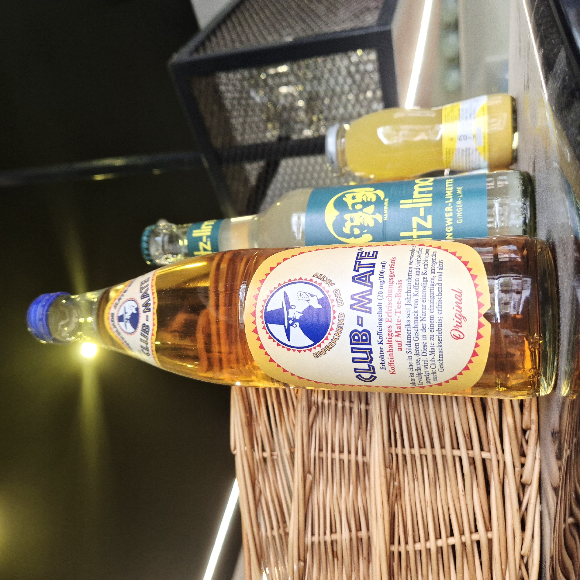 The image shows three bottles of beverages. The foremost bottle is a Club-Mate drink, characterized by its golden liquid and blue cap. Its label is predominantly yellow with a blue circle containing a man wearing a hat. The brand name is clearly visible, along with the word "Original." Behind the Club-Mate is a bottle of Fritz-Limo with a blue label indicating "Ingwer-Limette" (Ginger-Lime) and "Hamburg." A third bottle, containing a yellow liquid, is situated further back. The bottles rest on a wicker basket. The background is blurry, with glimpses of what appears to be a metallic structure. The text on the bottles is in German. The image does not show any location or reference to Strasbourg, France.