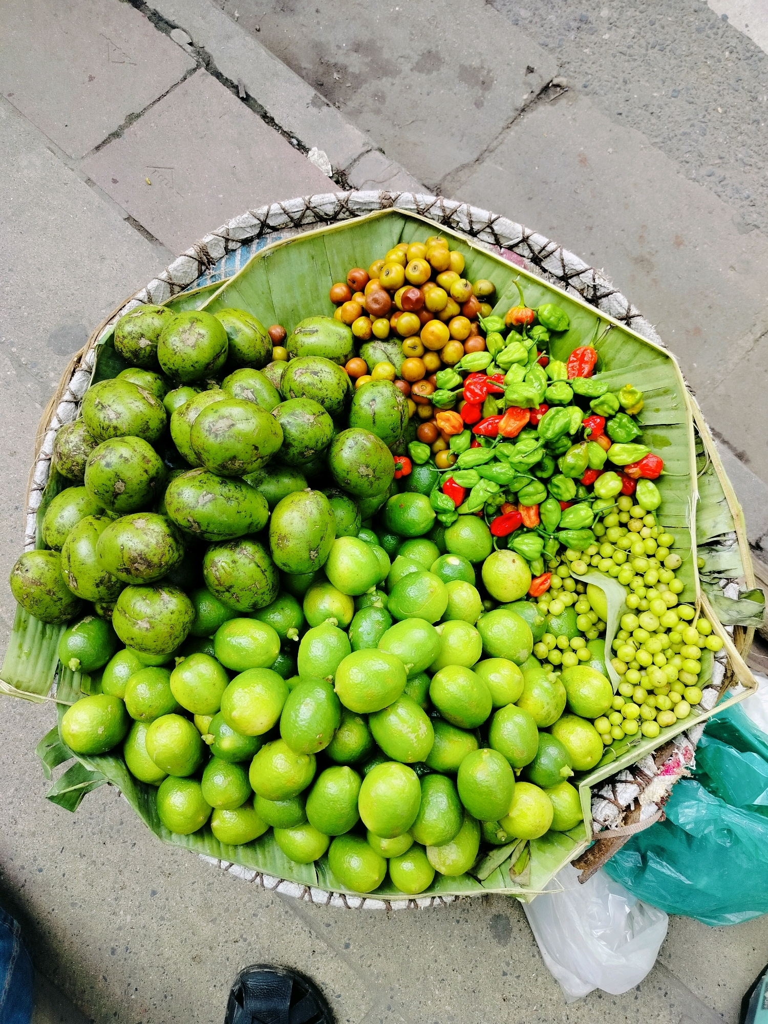 A high-angle medium shot captures a fruit vendor's stall in Dhaka, Bangladesh, displaying a colorful assortment of produce arranged in a woven basket lined with banana leaves. The basket is filled with a variety of fruits and vegetables, including large green fruits that resemble avocados, smaller green fruits that appear to be limes or small mangoes, a pile of tiny green berries, and a mix of red and green small peppers. The fruits are tightly packed, creating a vibrant mosaic of green, yellow, and red hues. The stall is situated on a paved city street, with cracked concrete visible in the background, hinting at a bustling urban environment. Part of a person's foot in a sandal is visible at the bottom right of the frame, and a crumpled plastic bag is also present, suggesting an active marketplace. The overall impression is one of a lively street scene in a South Asian city, highlighting the fresh produce available to shoppers.