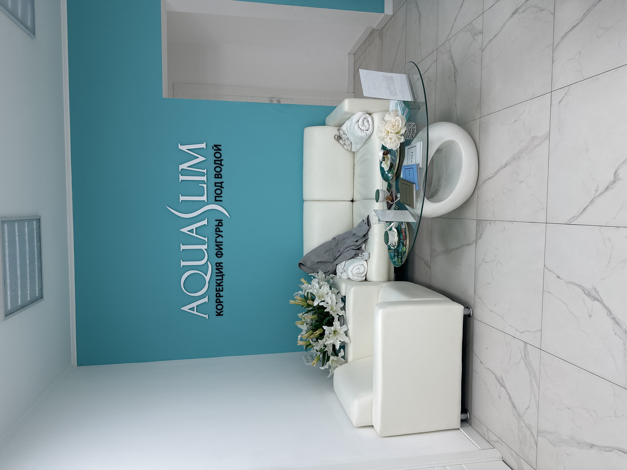 This image captures a modern, unoccupied waiting area within the AQUASLIM wellness center, identified by its logo "AQUASLIM КОРРЕКЦИЯ ФИГУРЫ ПОД ВОДОЙ" (AQUASLIM Body Contouring Under Water) on a prominent teal accent wall. This facility is located at Presnenskaya Naberezhnaya, 10с2, Moscow, Russia, within the Moscow City complex.
The room features an L-shaped white upholstered sofa unit on a white tiled floor with grey veining. A grey throw blanket and two rolled white towels are arranged on the sofa. A large bouquet of white lilies rests on the corner of the seating unit. In front of the sofa is a white, oval-based glass coffee table. On the table, two light green teacups with saucers, a plate containing small pastries, a small arrangement of white decorative flowers in a silver vase, and multiple fashion-themed books (including "Gucci" and "Dior") are displayed. Two clear acrylic stands with printed informational materials are also present on the table. The ceiling is white and features recessed rectangular fluorescent light fixtures. A white door frame is visible on the right side of the image. No individuals are present in the scene.