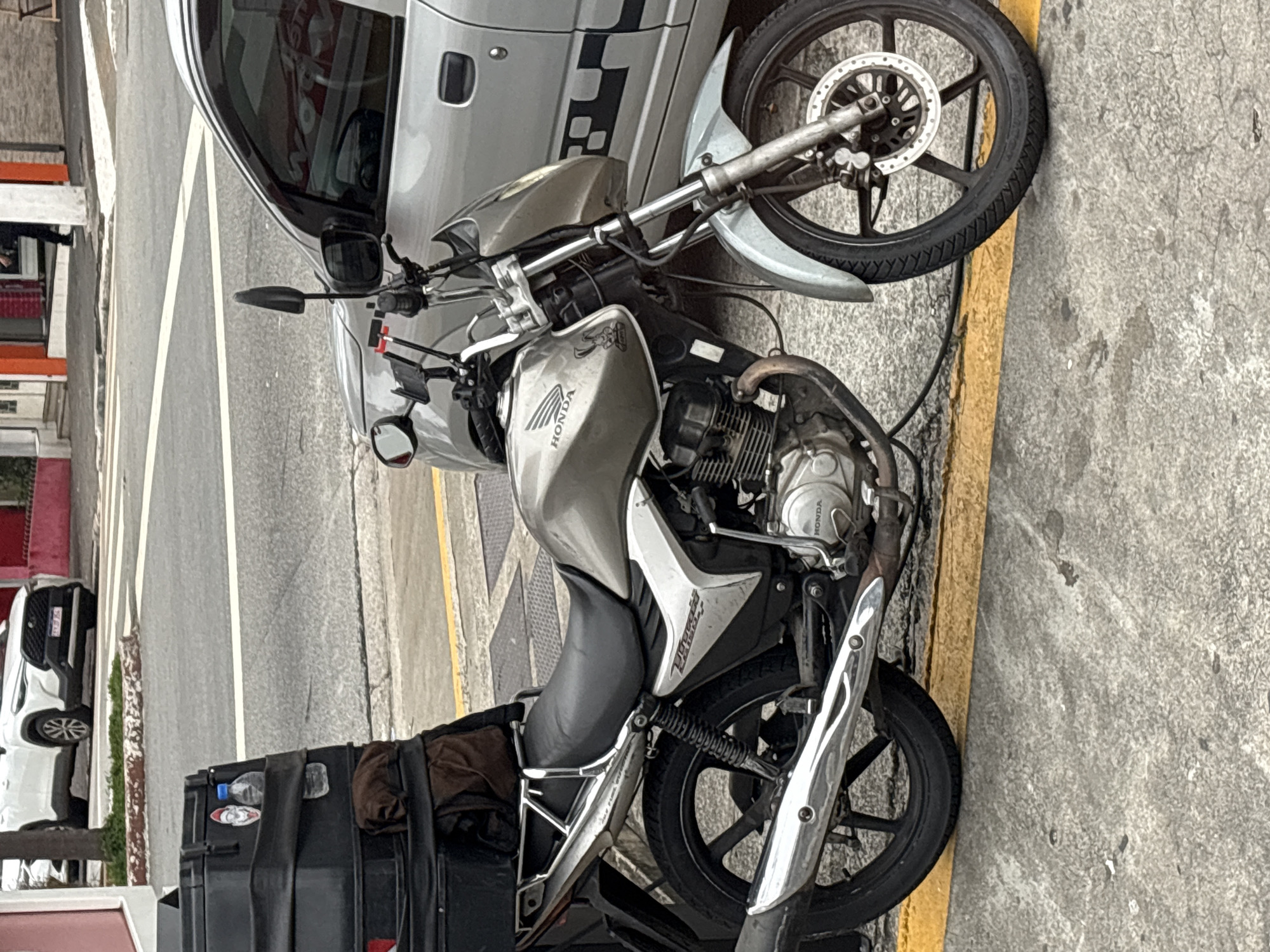 A silver Honda motorcycle is parked on the right side of the image, close to a silver car. The motorcycle has a black storage box attached to its rear, containing a brown cloth. The motorcycle is on a paved street with a yellow line marking a lane. In the background, a white SUV is parked, and buildings with red and orange facades are visible. The scene appears to be a street in a city, potentially in São Paulo, Brazil, given the style of the buildings and vehicles. There are no people visible in the frame.