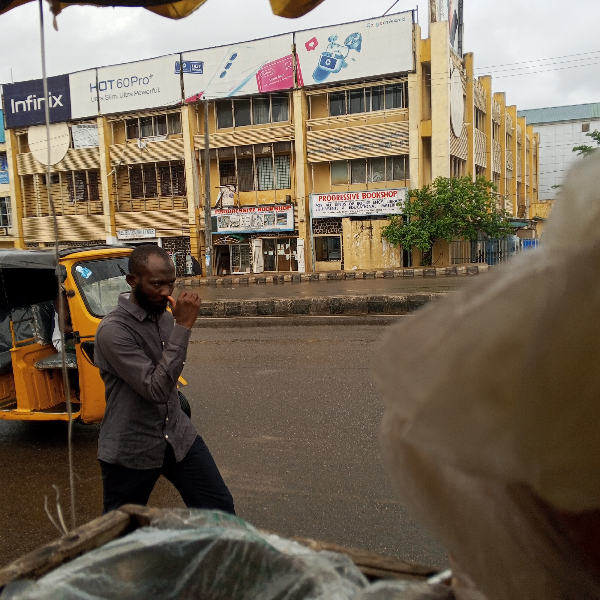 In Maiburiji, Nigeria, a man with a beard, wearing a dark gray collared shirt and dark pants, stands next to a yellow auto-rickshaw. He is looking down, with his right hand raised to his mouth, appearing to be contemplating or perhaps chewing something. The auto-rickshaw has a black canopy and a grey seat. In the background, a multi-story building with yellow facade features large advertisements on its upper levels. One prominent advertisement displays "Infinix HOT 60 Pro+" and "Ultra Slim. Ultra Powerful." Another advertisement features "Google on Android" and images of earbuds and a phone. On the ground floor of the building, there are two shopfronts labeled "PROGRESSIVE BOOKSHOP." The scene is outdoors, on a paved road that appears wet, suggesting recent rain. The sky is overcast. The overall impression is that of a typical urban street scene in Nigeria.