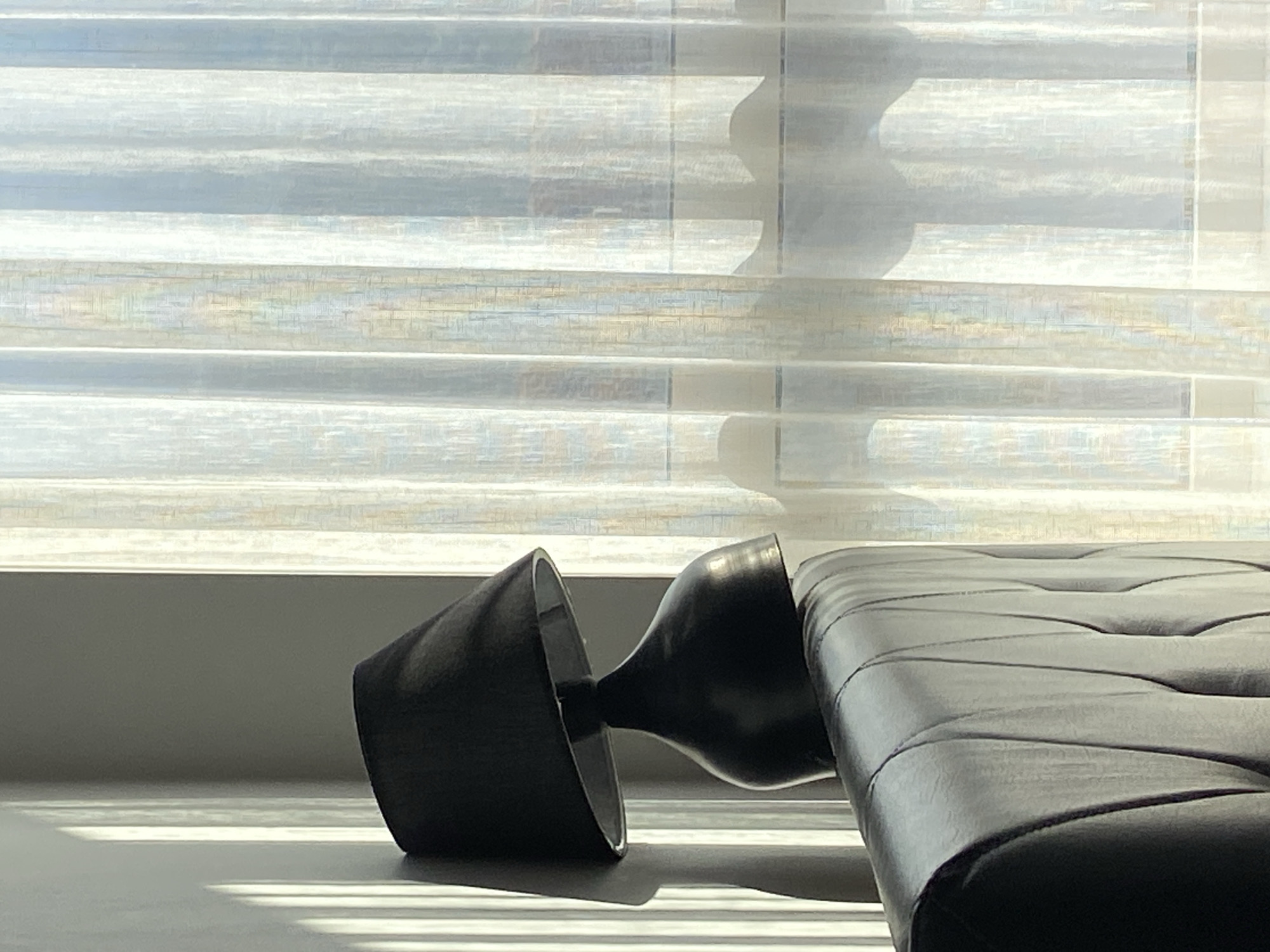The image captures an indoor scene bathed in natural light streaming through a window. In the lower left, a distinctive black, two-part object rests on a bright, horizontally striped surface, likely a windowsill or table. One part of the object resembles a wide, open cone lying on its side, while the other is a more rounded, bulbous shape, connected to the first. To the right, a section of black, tufted or quilted leather furniture, possibly an armchair or sofa armrest, is partially visible.
The background is dominated by horizontal window blinds, which appear semi-sheer or translucent, diffusing the bright sunlight. Shadows of external elements, possibly architectural features or foliage, are cast onto the blinds as soft, undulating shapes. The light coming through the blinds creates a striking pattern of bright stripes and distinct shadows on the surface in the foreground. This strong, directional lighting indicates it is daytime. No people or activities are present, and no text is visible within the frame. The overall atmosphere is quiet and still. While the location context is provided as Budva, Montenegro, there are no specific visual cues in the image itself to confirm this.