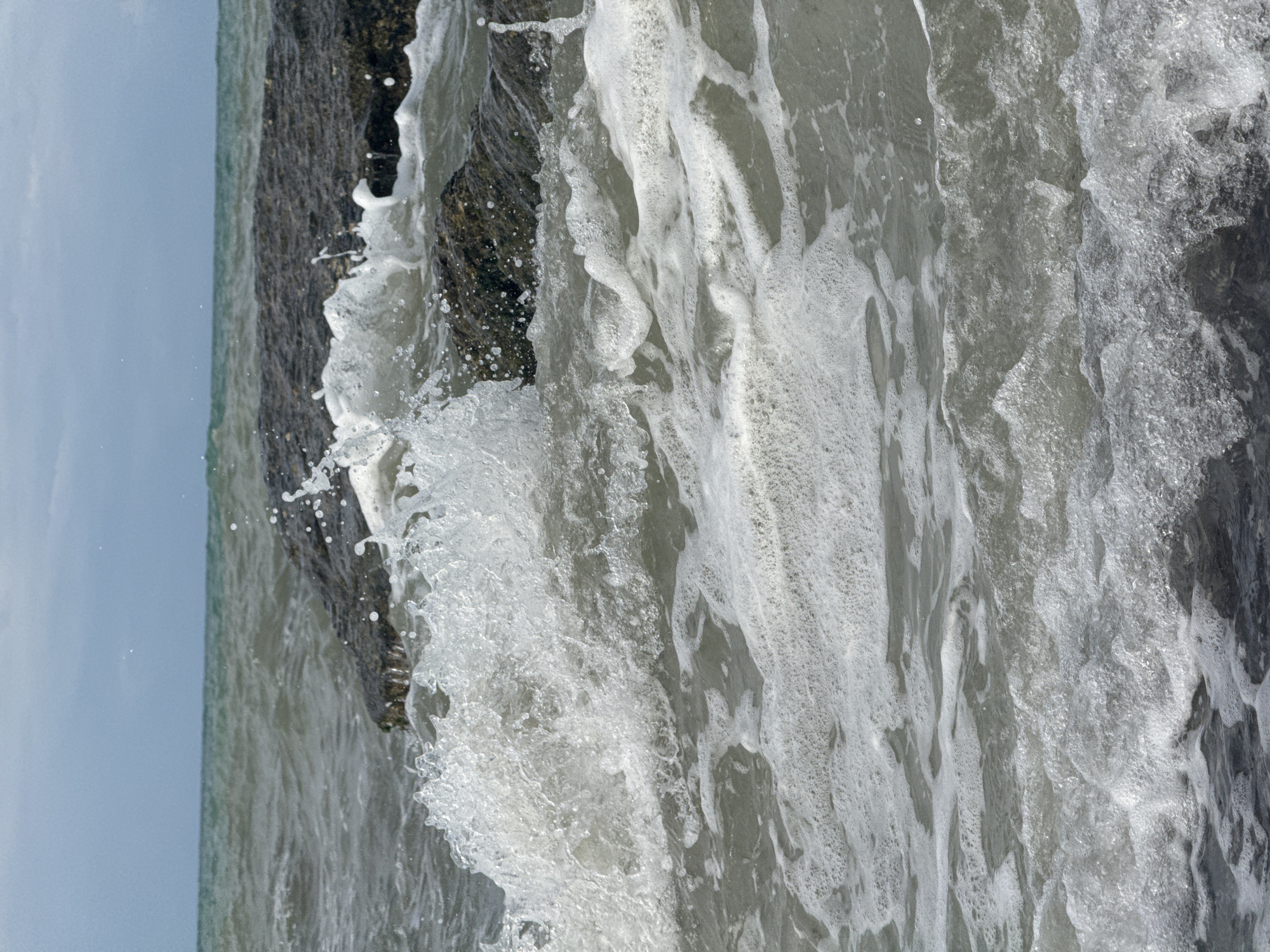 The image shows a close-up of waves crashing against dark rocks in the sea. The water is turbulent and frothy, with white foam and spray. The rocks are dark and jagged, providing a stark contrast to the white water. The sea stretches out to the horizon, where it meets a hazy sky. The scene is natural and unpopulated, with no people or man-made objects visible. The location is the Caspian Sea coast near Aktau, Kazakhstan, characterized by its rocky shorelines and often choppy waters, particularly during overcast weather as suggested by the sky in the image.