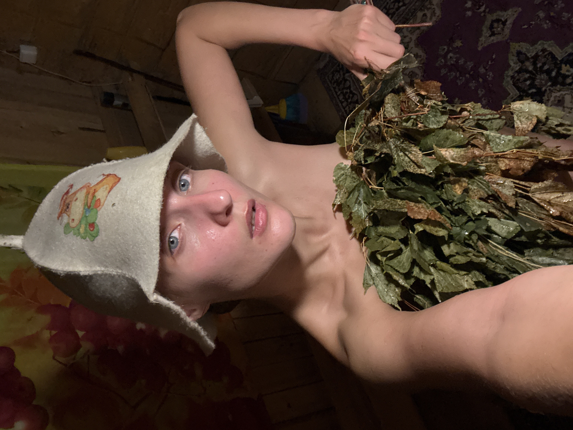 The image shows a person wearing a white felt hat with an image of a cartoon character on it. They are holding a bundle of green and brown leaves. The person has light skin and blue eyes, and appears to be sweating. The setting appears to be a wooden room, possibly a sauna, with a patterned rug visible in the background. The lighting is dim, suggesting it might be nighttime or the interior of a building. The person's expression is unreadable due to the angle. There is no visible text in the image. The context provided indicates the location is Sharkan, Russia.
