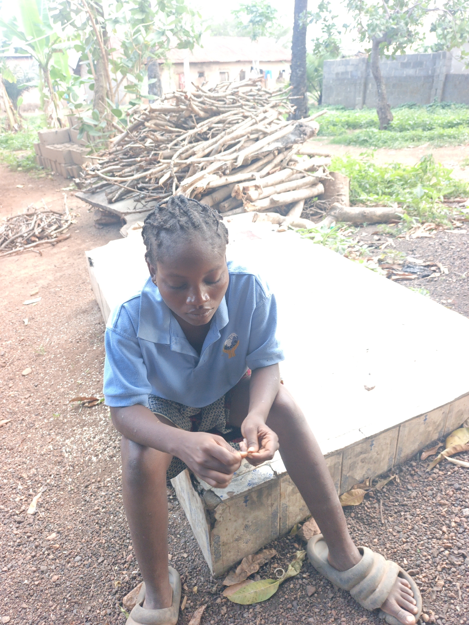 A young person with braided hair sits on a concrete structure outdoors. They are wearing a blue collared shirt, patterned shorts, and slides. The person is looking down and appears to be manipulating something small in their hands. Behind the person is a large pile of branches and logs. The ground is reddish brown gravel. In the background are trees, vegetation, and buildings. The scene is likely in Akwanga, Nigeria.