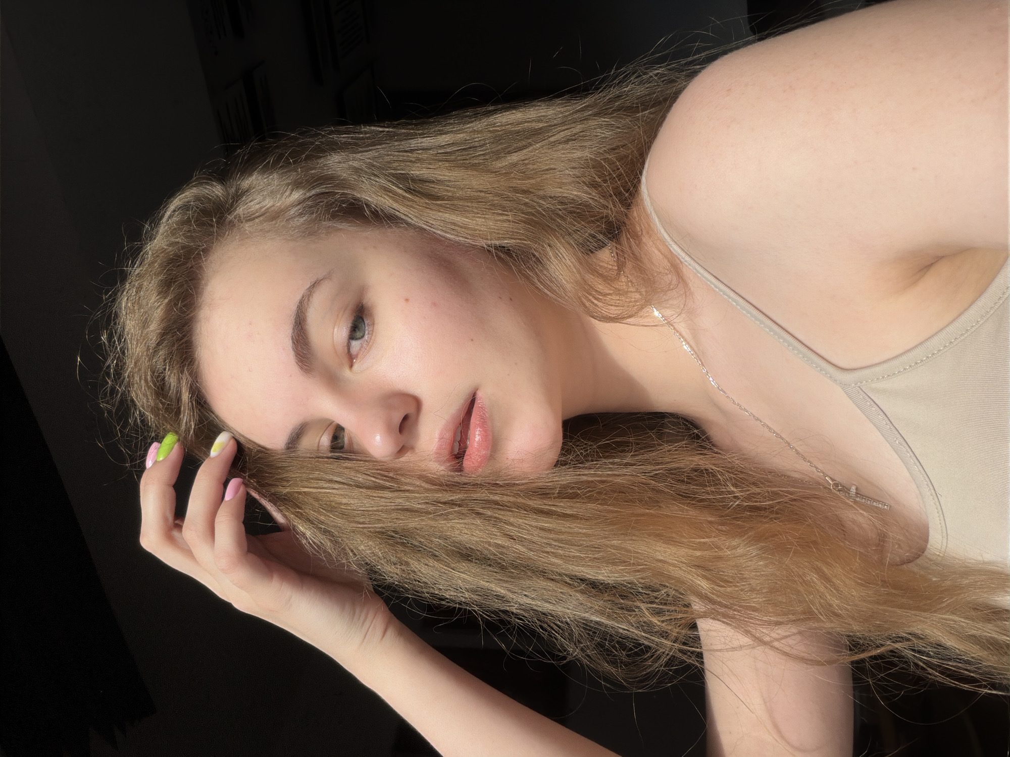 The image is a close-up, self-portrait of a young woman with blonde hair and light skin. She is looking towards the camera with a neutral expression. Her left hand is raised, touching her hair. She is wearing a light-colored tank top and a silver necklace with a cross pendant. The lighting suggests she is indoors near a window, with sunlight casting shadows. There is no identifiable information about her location in Dnipro, Ukraine, or any specific environmental details that would place her in a city. The background is dark and out of focus.