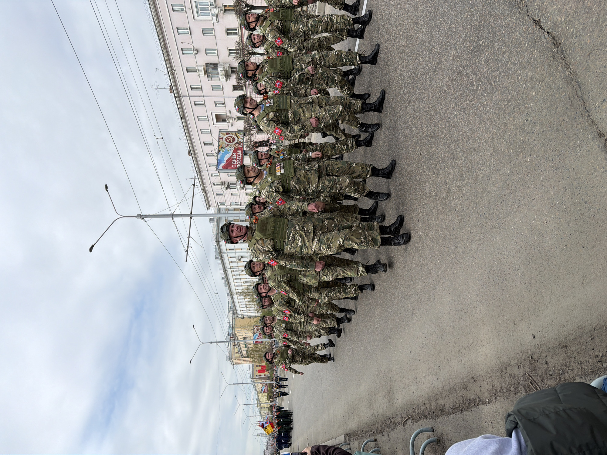 A parade of soldiers in camouflage uniforms marches down a city street in Krasnoyarsk, Russia. The soldiers are aligned in neat rows. Each soldier wears a helmet and carries gear. A building with multiple windows is visible on the left side of the street. Streetlights line the street. Banners are visible amongst the marchers, including one with the text "С Днем Победы" (Happy Victory Day). In the foreground, a person's arm and clothing suggest they are watching the parade.