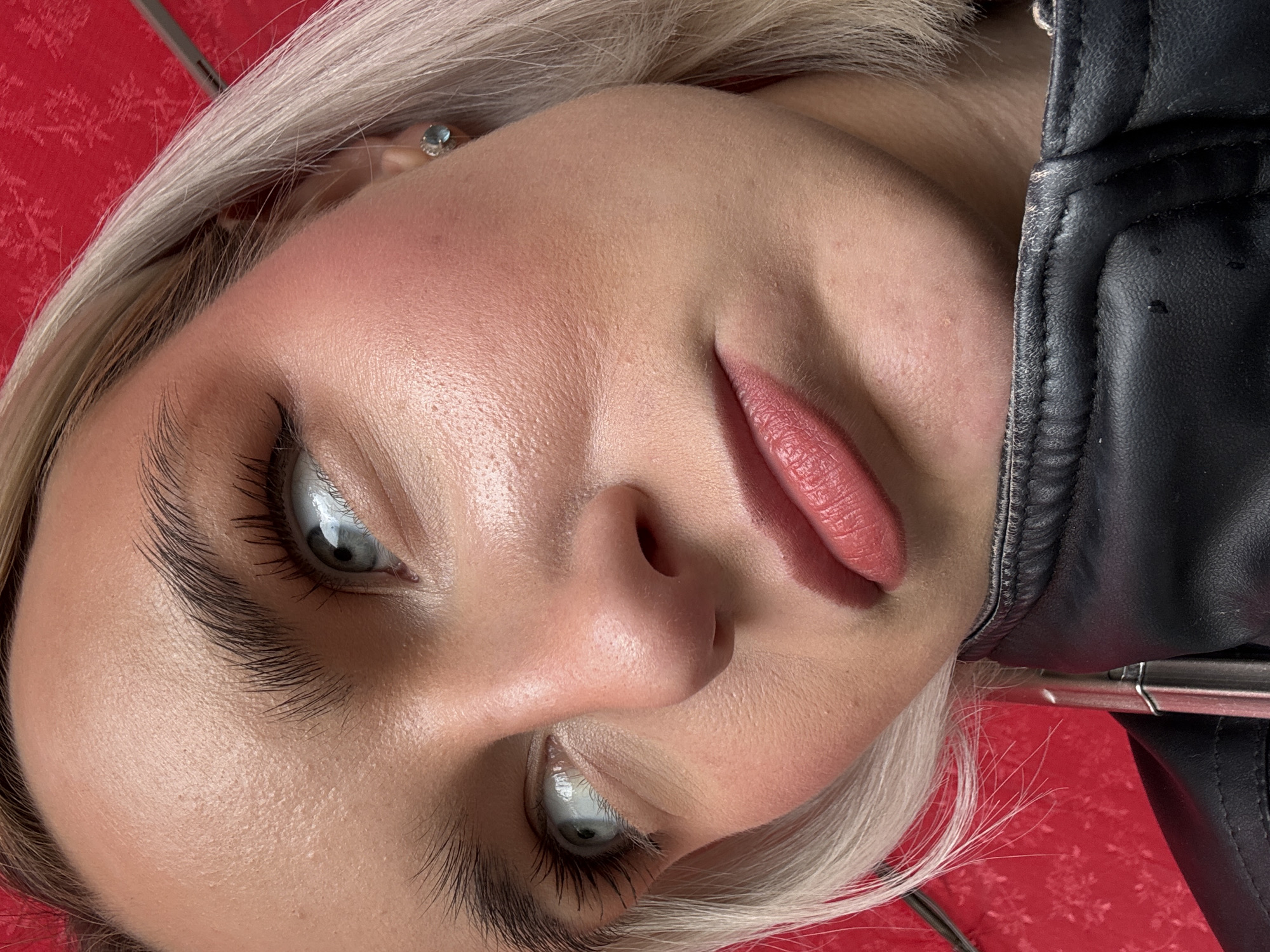 A close-up, upward-facing shot shows a woman with light blonde hair and greyish-blue eyes. She has fair skin, full lips, and dark, defined eyebrows. She is wearing a black leather jacket. The background is a blurry red with a subtle pattern. An earring is visible in her ear. The scene does not include details that would allow identification of a location in Saint Petersburg, Russia.