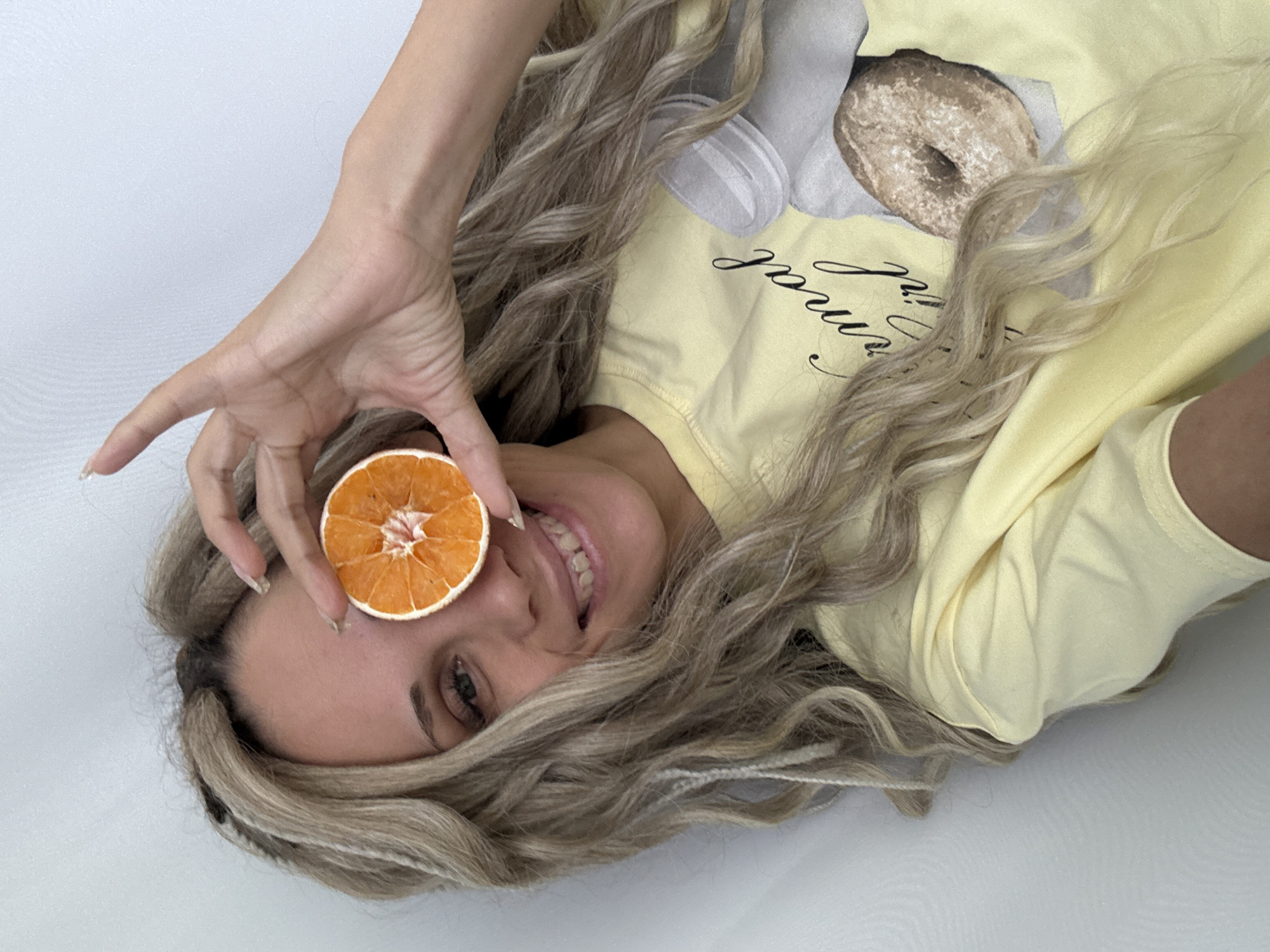 A woman lies on her side, holding a dried orange slice up to her eye. She is smiling and wearing a light yellow shirt with a donut graphic. Her hair is long and blonde, with dark roots visible. The background is a plain white surface. The location is not discernible from the image.