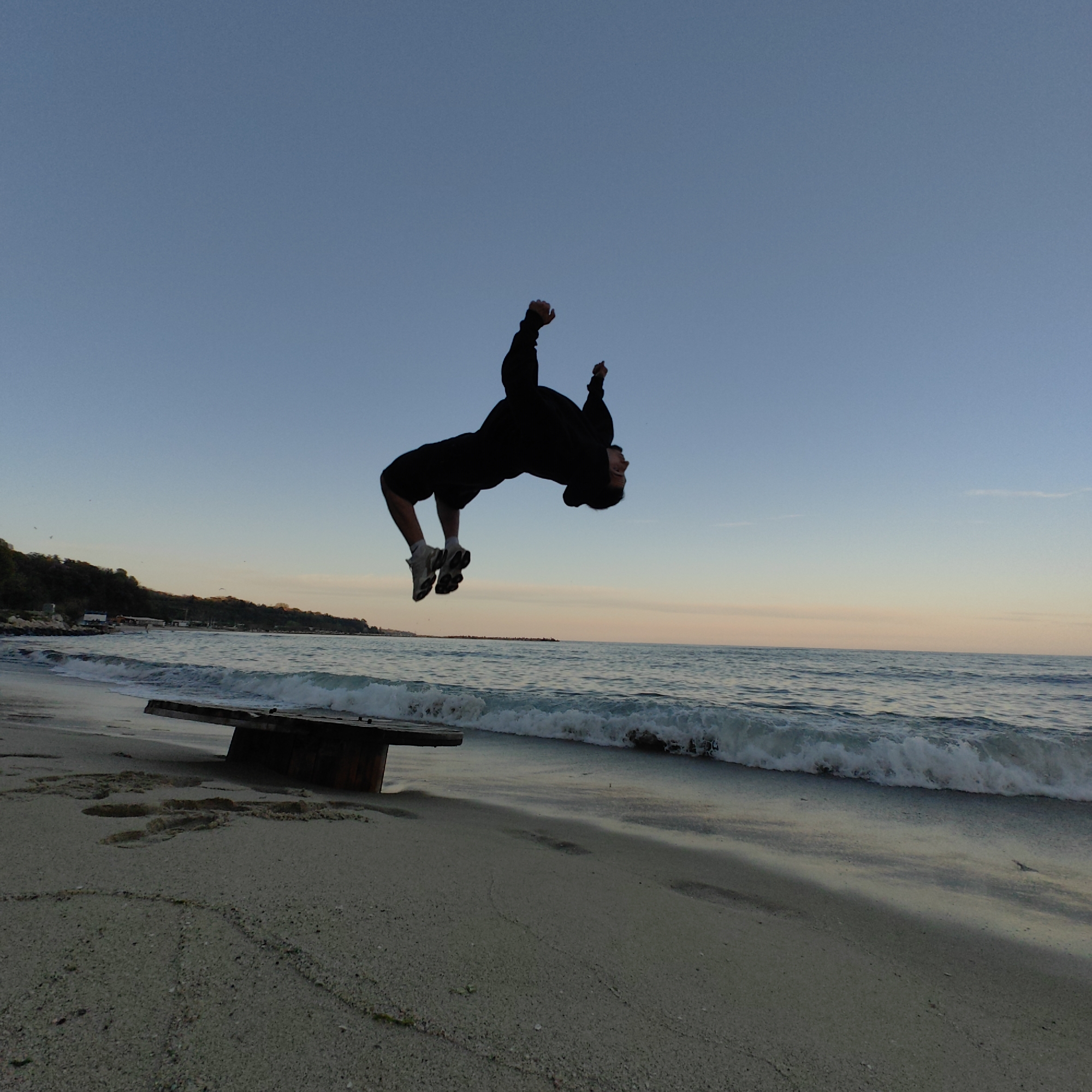A person in dark clothing is performing a backflip on a beach. The person is mid-air, with their body inverted. The beach is sandy, with waves lapping the shore. In the background, there is a low-lying coastal area with some vegetation. A wooden structure is visible near the shoreline. The sky is clear with a gradient of colors from blue to pink near the horizon, indicating dawn or dusk. The location is likely a beach in Varna, Bulgaria.