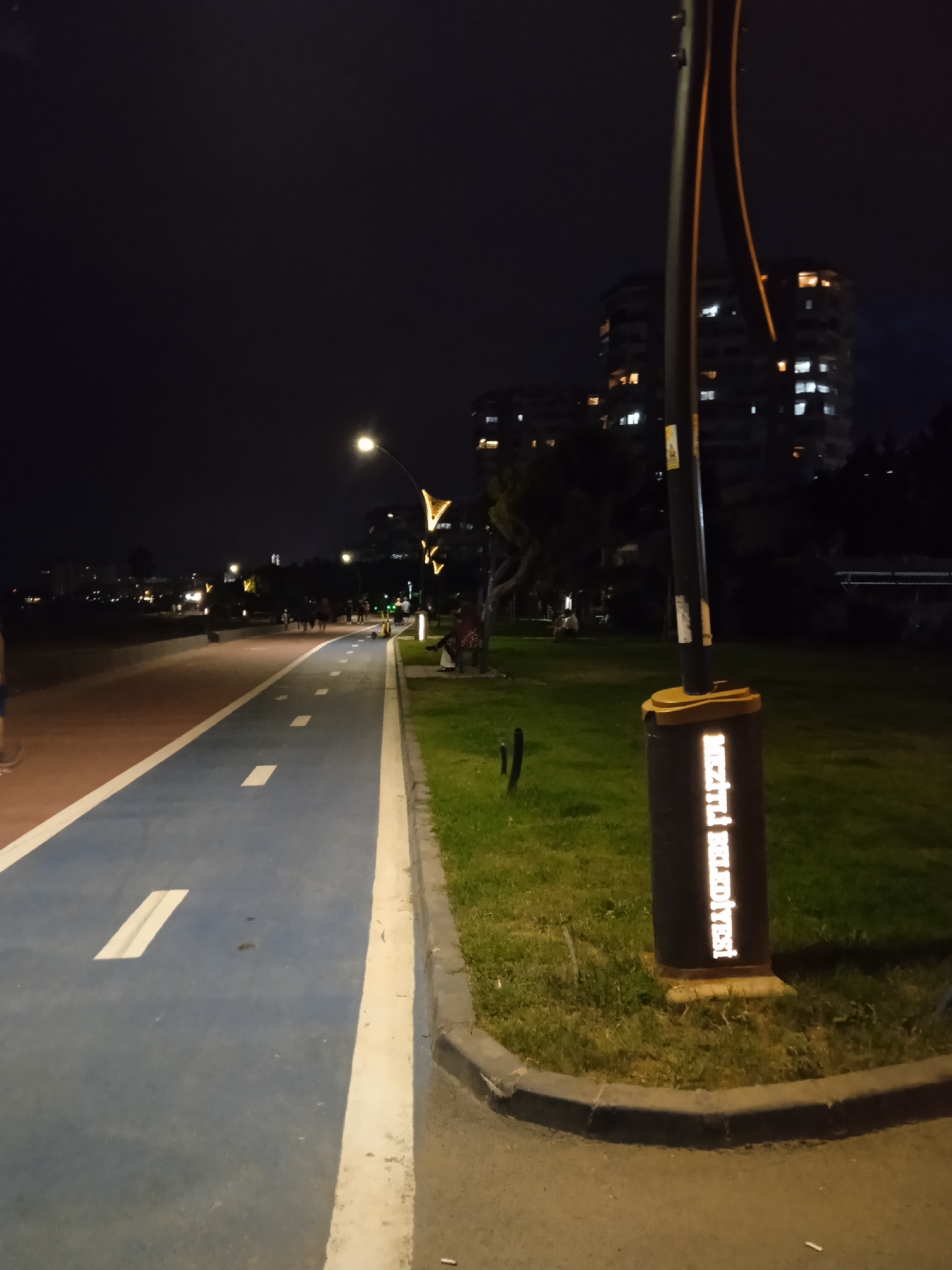 The image depicts a nighttime scene along a coastal promenade in Mezitli, Turkey. A blue and red asphalt path, divided by white lines, curves into the distance, suggesting a dedicated lane for cycling or jogging. Adjacent to the path is a well-maintained grassy area, bordered by a concrete curb. A tall lamppost stands prominently in the foreground, its base adorned with a cylindrical bin that features reflective lettering reading "Mezitli Belediyesi" (Mezitli Municipality). In the background, illuminated apartment buildings with visible lights suggest an urban setting. Several individuals are casually walking along the path and sitting on benches in the grassy area, enjoying the evening. The overall atmosphere is tranquil and urban, illuminated by artificial lighting.