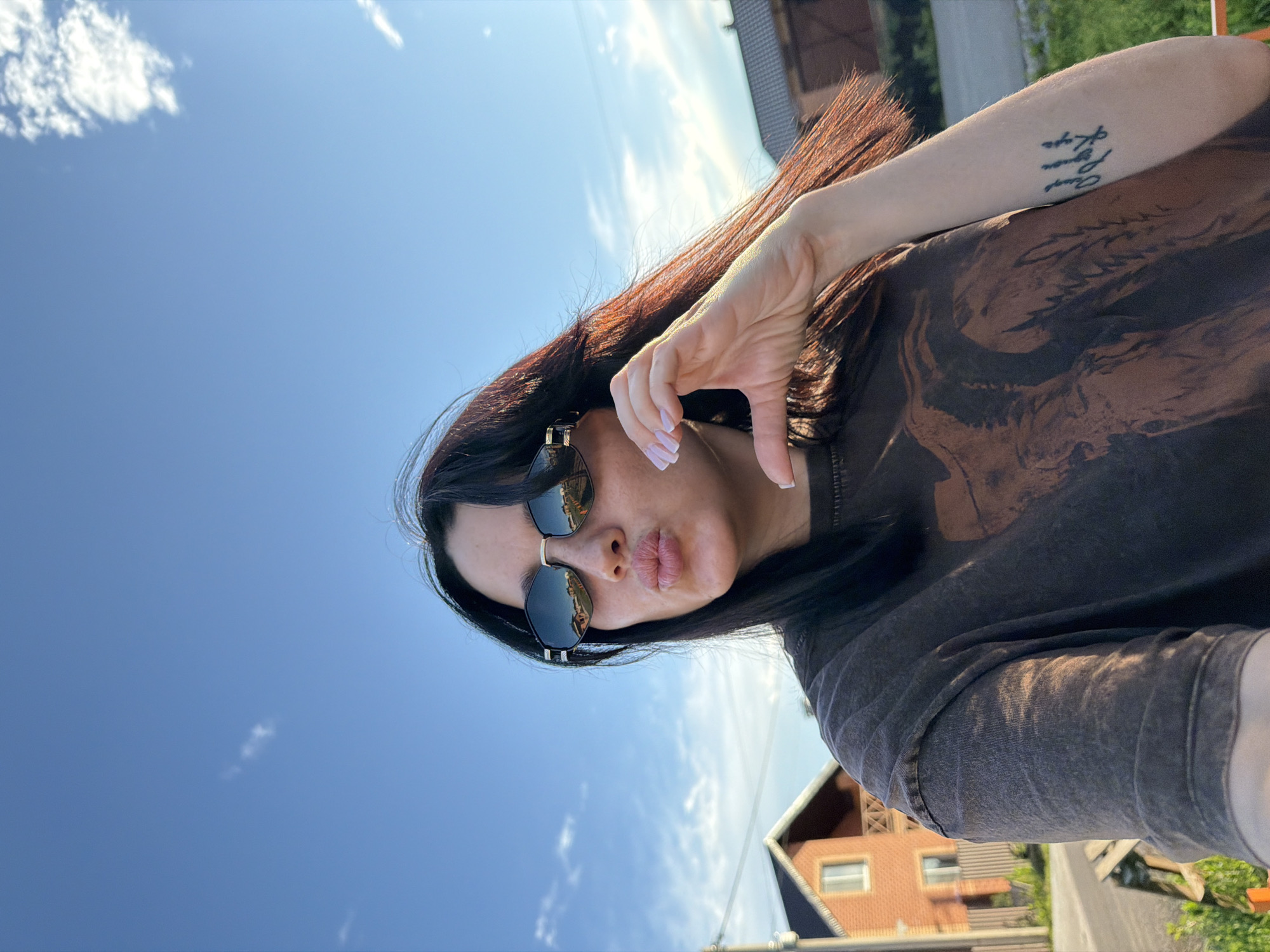 A young woman with long, dark hair and sunglasses is captured in a selfie outdoors. She is puckering her lips and forming a heart shape with her fingers, her hand resting near her chin. She is wearing a dark, possibly brown, t-shirt with a graphic print. A tattoo is visible on her left forearm. In the background, a red brick house with a wooden fence and a glimpse of a rural road can be seen, suggesting a location in a suburban or rural area. The sky is bright blue with scattered clouds. The overall scene is casual and appears to be taken during daylight hours.