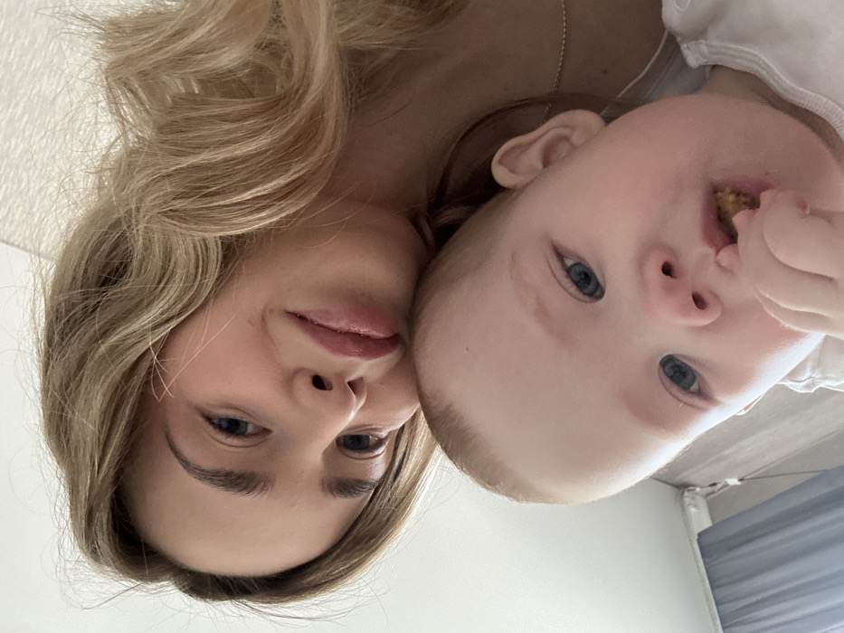 The image displays a close-up, high-angle interior shot featuring an adult female and an infant, situated in the city of Chaikovsky, Russia. The adult female, positioned in the upper frame, has light blonde, wavy hair and blue eyes, with a subtle smile. A thin, silver-colored chain necklace is visible on her neck. Directly below her, in the foreground, is an infant with light reddish-blonde hair and blue eyes. The infant is holding a small, textured, brownish object, consistent with a food item, in their right hand and is in the process of bringing it towards their mouth. Both individuals are partially clothed in white garments. The background consists of a white ceiling or wall, a visible section of a light blue or gray curtain on the left, and a white, horizontal fixture positioned above the curtain.