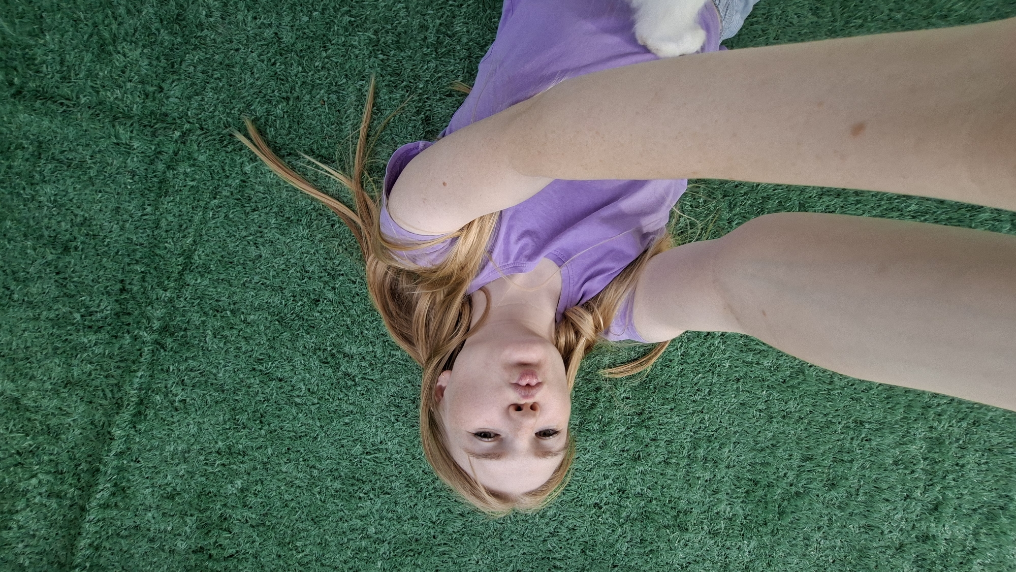 A young Caucasian girl with long blonde hair is lying on her back on green Astroturf. She is wearing a purple t-shirt and her arms are extended above her head, as if taking a selfie. Her lips are puckered, and her eyes are open. The image is taken from a high angle, looking down at the girl. The background is a uniform green Astroturf. The girl is positioned in the center of the frame, with her head towards the bottom and her feet towards the top. There is a small white object, possibly a toy or a pet, resting on her chest, partially obscured by her arms. There are no discernable landmarks or clues that would indicate a specific location in the city of Большой Букор, Russia.