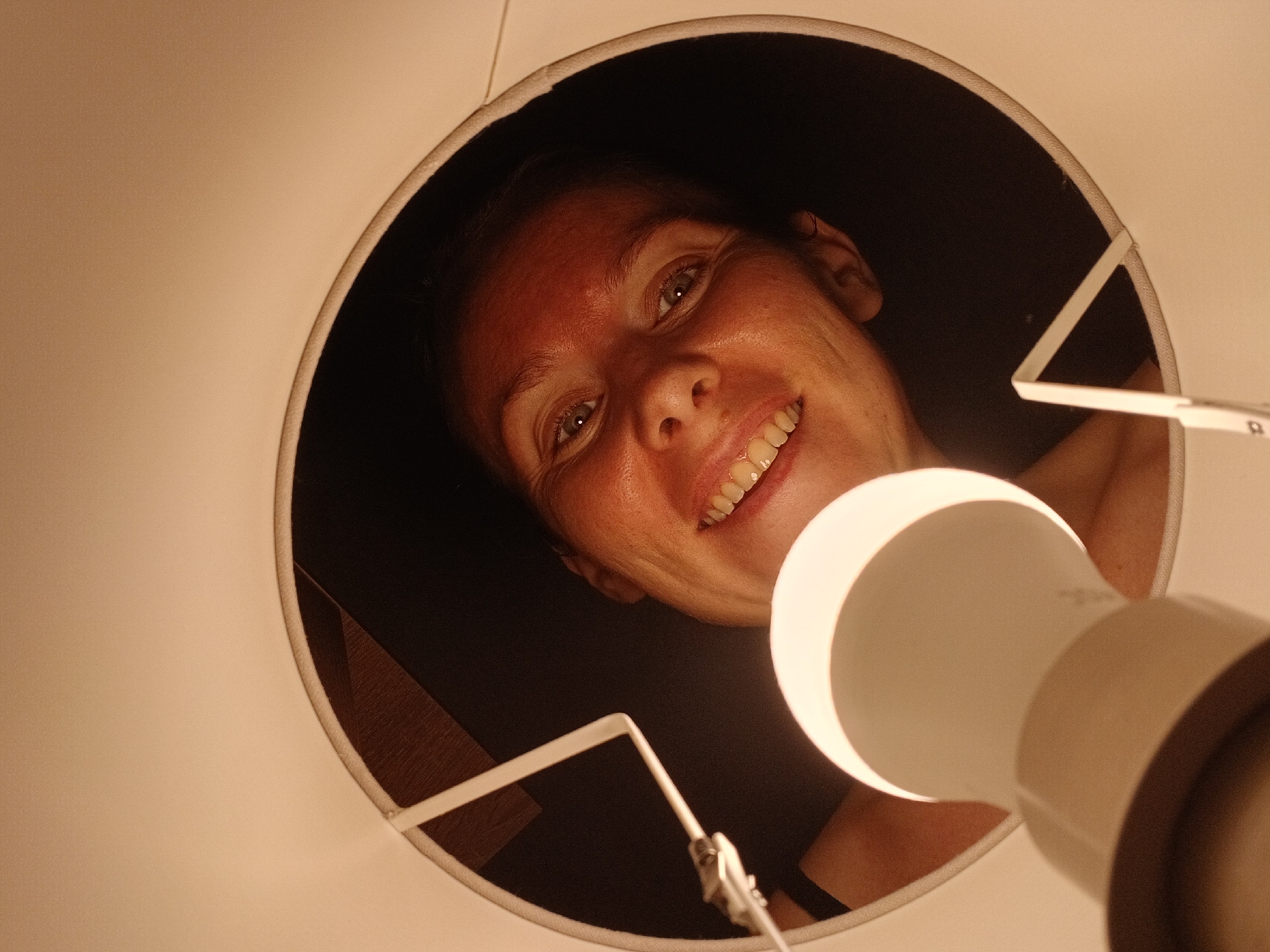 This is a close-up, low-angle shot of a woman's face looking up from inside a lampshade. The woman has light skin, blonde hair, and is smiling, showing her teeth. Her eyes are open and she appears to be looking directly at the camera. The lighting is warm and suggests an indoor setting, possibly at night, given the prominence of the lamp. The lampshade is white and circular, framing the woman's face. The image is focused on the woman and the lamp bulb. There is no identifiable information in the image that would allow for its location to be determined as Yekaterinburg, Russia.