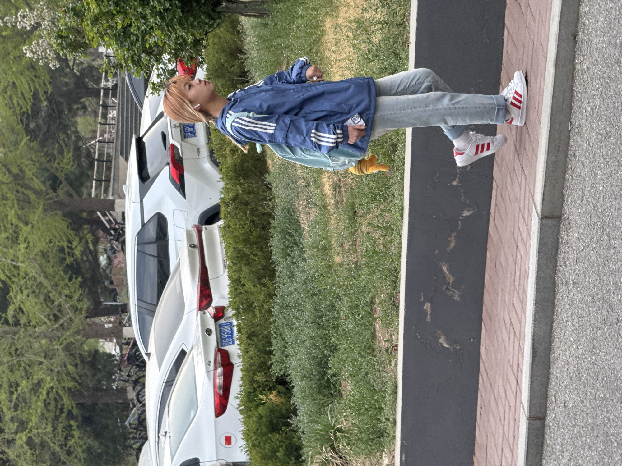 A person is standing on a sidewalk in Zhongtun, China, adjacent to a street with parked cars. The person is wearing a blue jacket with white stripes, light-colored jeans, and white sneakers with red stripes. They are also wearing earbuds. The person has light-colored hair and is looking upwards. Next to the sidewalk is a strip of grass or greenery, and behind that are several white cars parked along the street. The license plates of the cars are visible. In the background, trees and other vegetation can be seen.