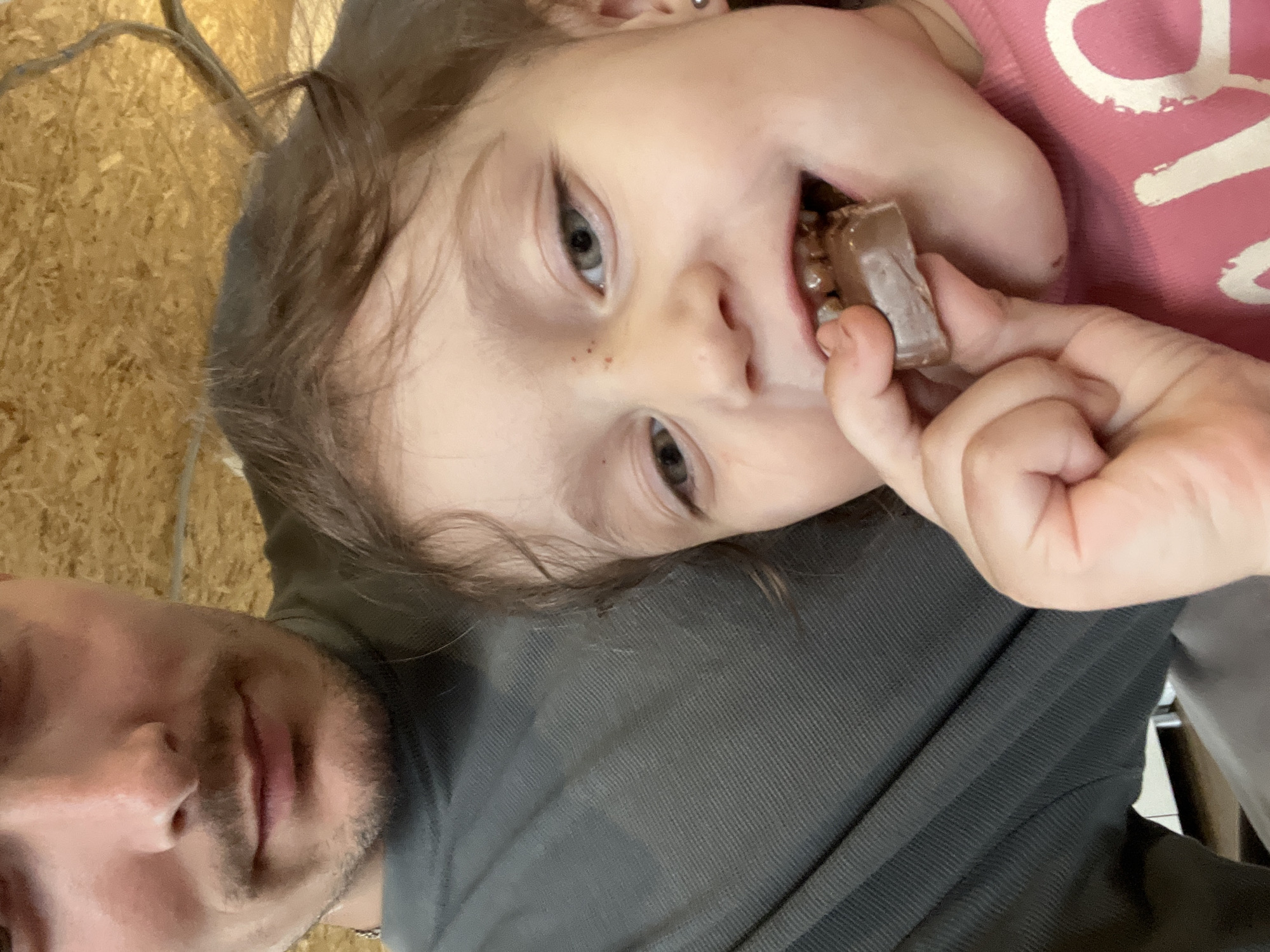 A selfie-style image shows a young child with light skin, brown hair, and grey eyes, eating a chocolate bar. The child is wearing a pink shirt with a white graphic. The child's head is tilted, and they are holding the chocolate bar in their right hand, bringing it to their mouth. A portion of an adult's face is visible in the lower-left corner, indicating they are holding the child. The adult has facial hair and is wearing a grey shirt. The background consists of a light brown wooden surface and a sparse tree branch, suggesting an indoor setting. There's no information to suggest the location is Chaikovsky, Russia.
