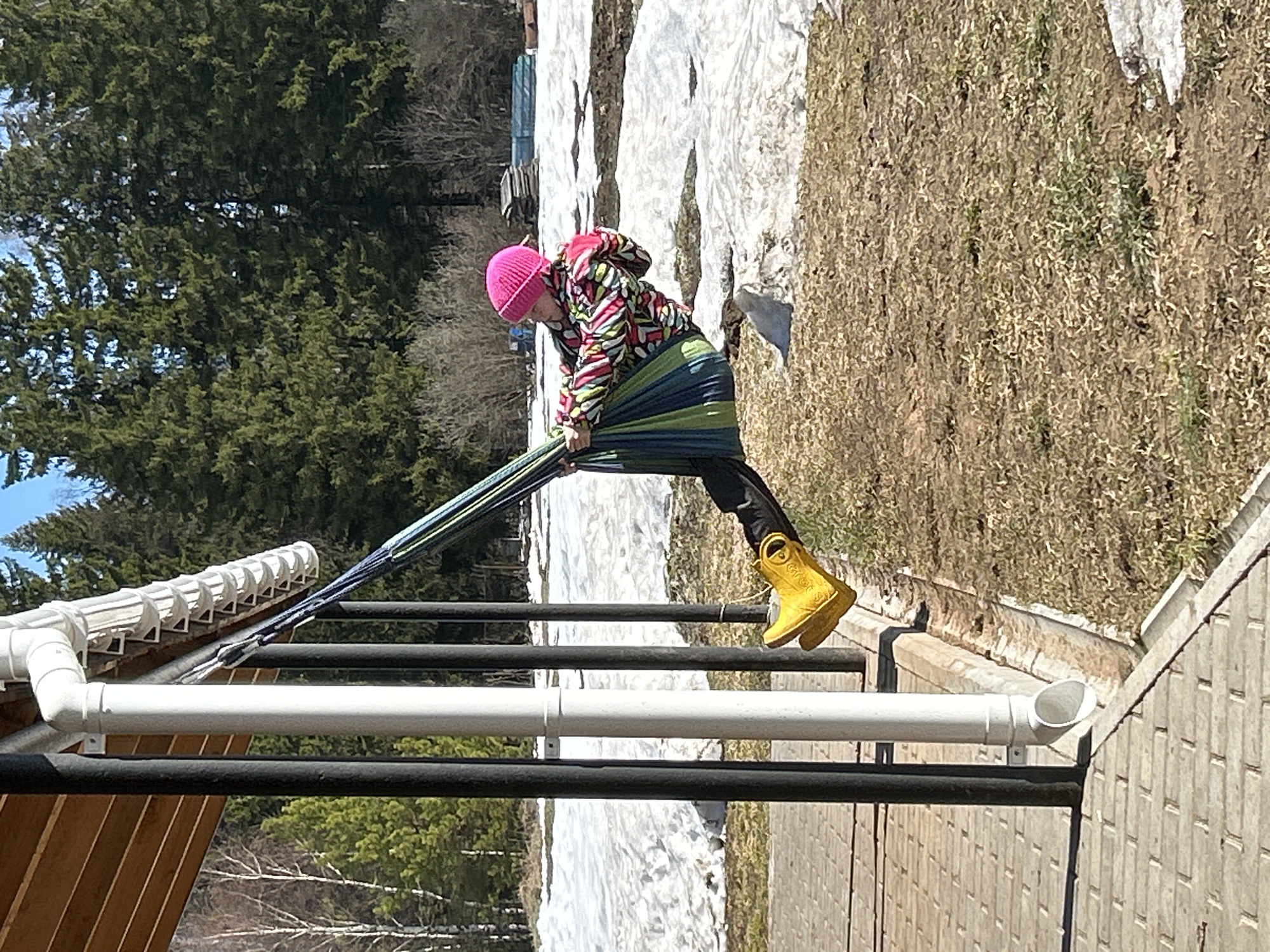 A young person wearing a pink hat, a colorful jacket, black leggings, and yellow boots is positioned in a hammock. The hammock appears to be stretched between a white drainage pipe and another structure, possibly a fence. The ground shows patches of snow and dried grass, with bare trees visible in the background along with a dense evergreen forest. The photograph was taken during daylight hours. The location is Уральское, Russia.
